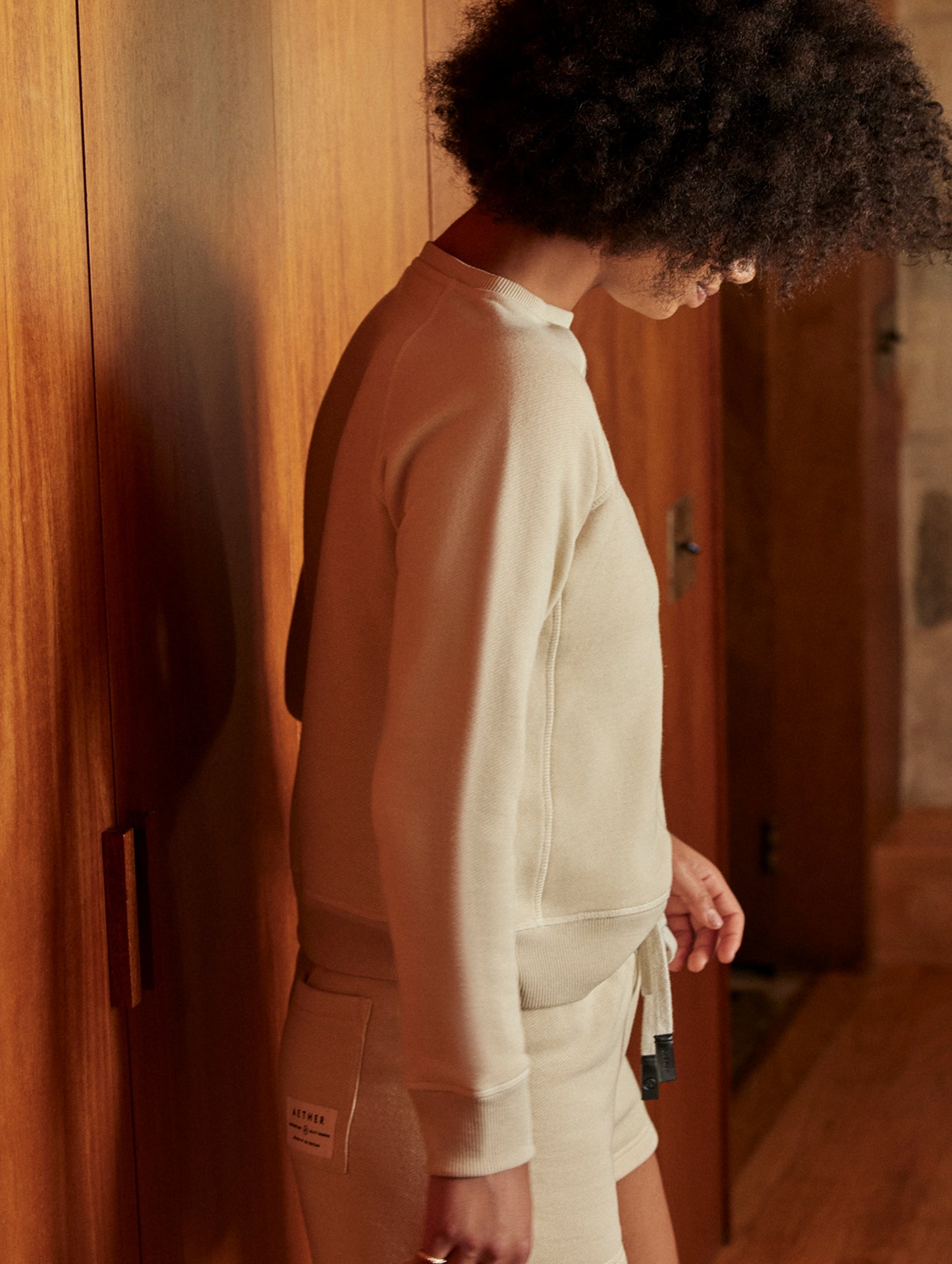 Side profile indoor lifestyle image of woman wearing Foundation Raglan Sweatshirt in Bone white from AETHER Apparel.