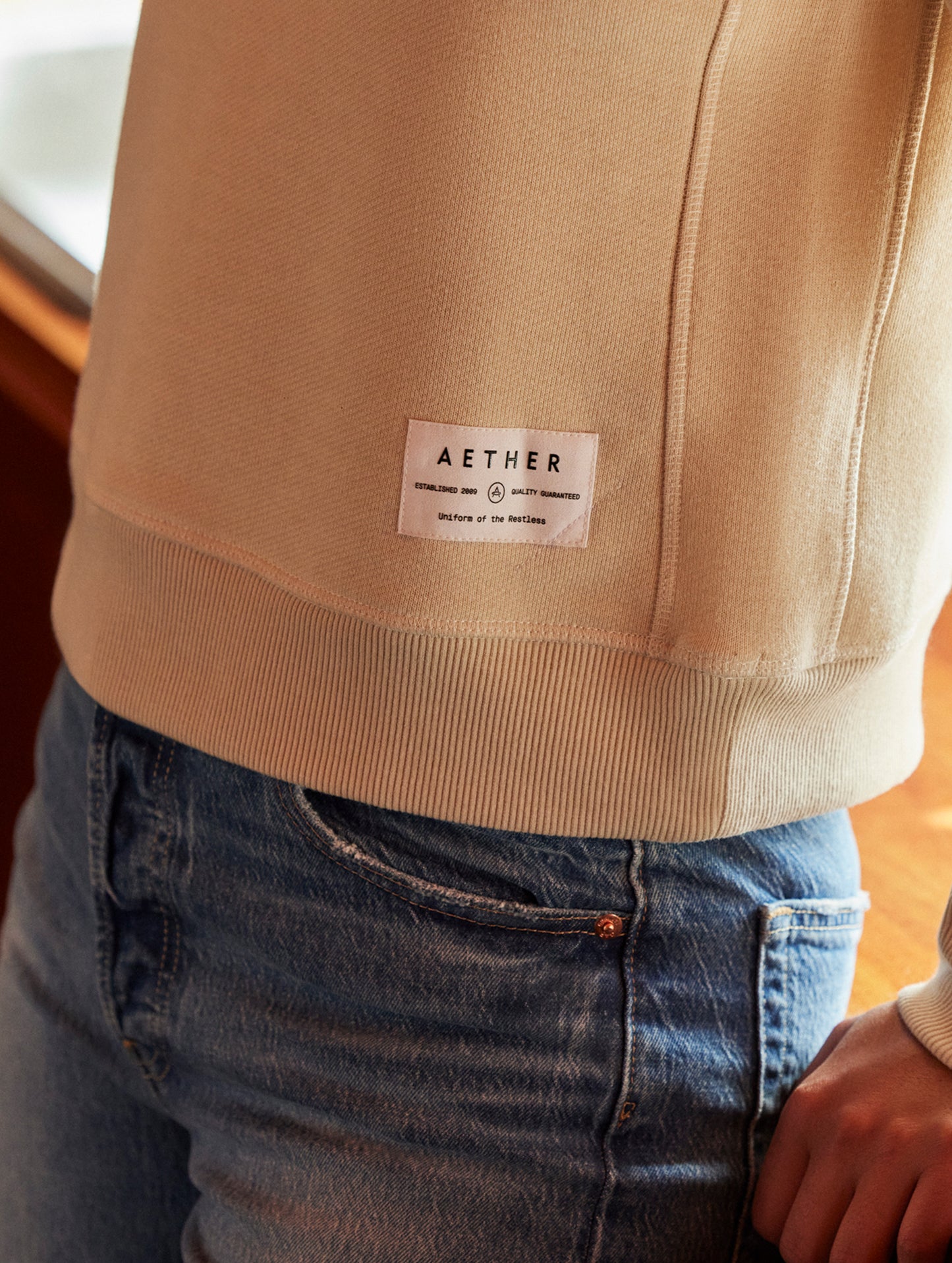 Closeup detailed view of elastic hem and logo patch on women's Foundation Raglan Sweatshirt in Bone white from AETHER Apparel.