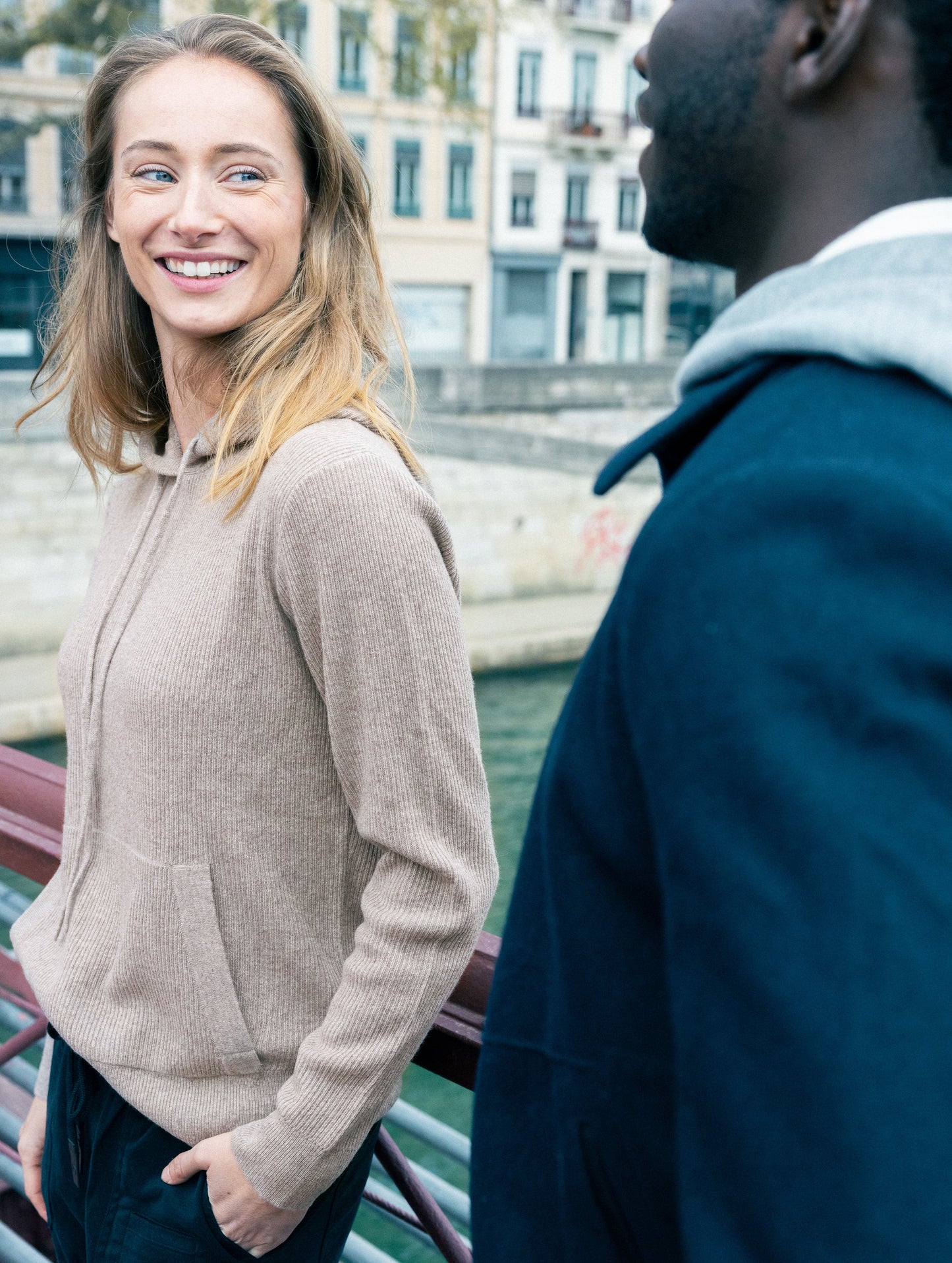 Outdoor lifestyle image of woman leaning against railing wearing Lawrence Cashmere Hooded Sweater in Toast Brown from AETHER Apparel.
