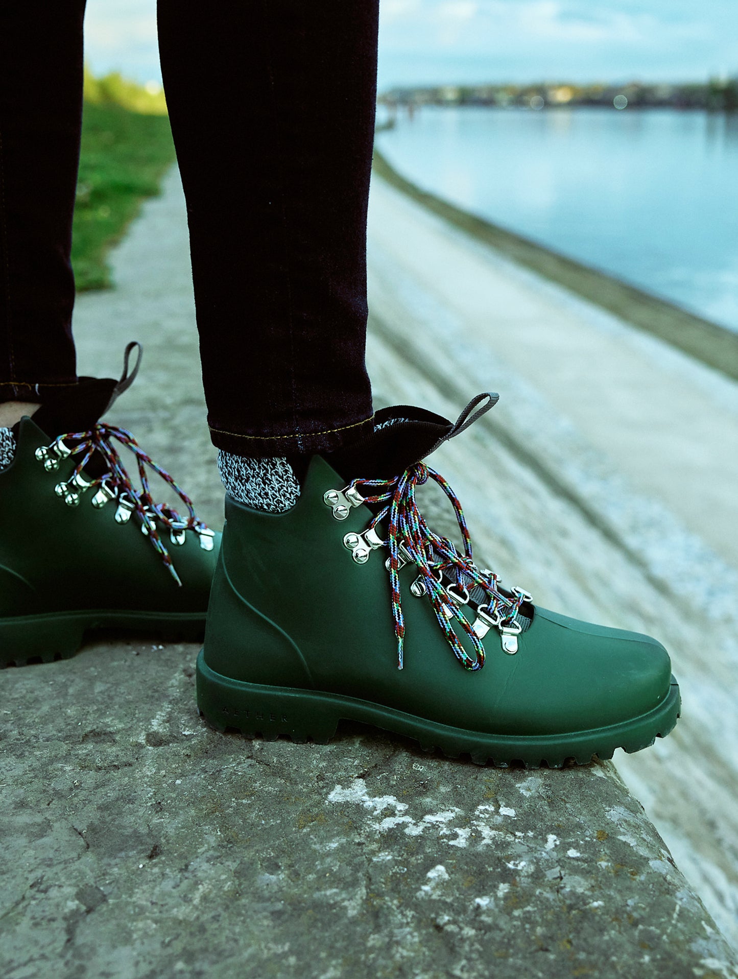 Outdoor lifestyle image of woman standing near body of water wearing Rain Boot in Dark Green from AETHER Apparel.