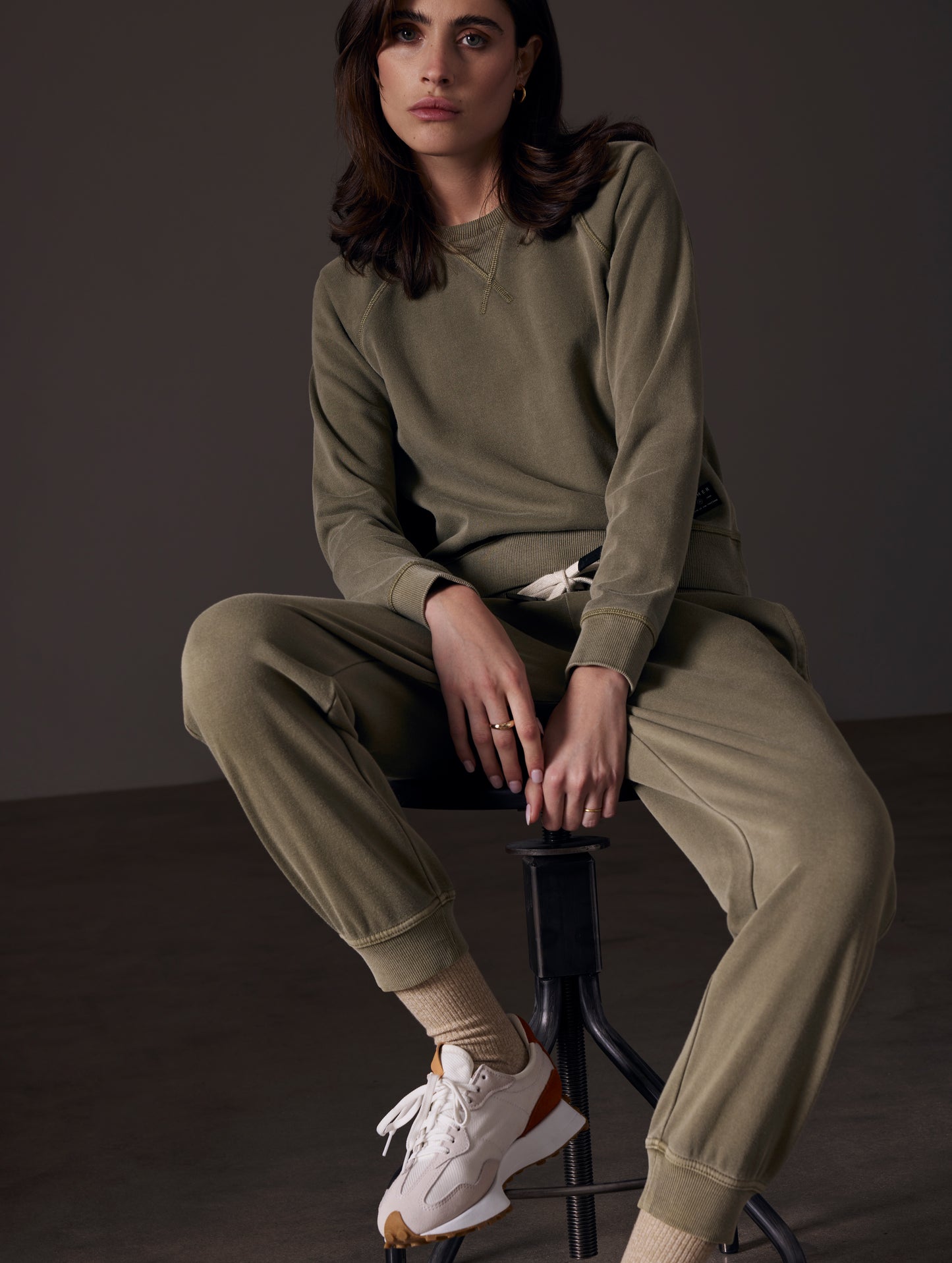 Front angled view of woman sitting on stool wearing Solstice Lightweight Raglan Pullover in Command Green from AETHER Apparel.