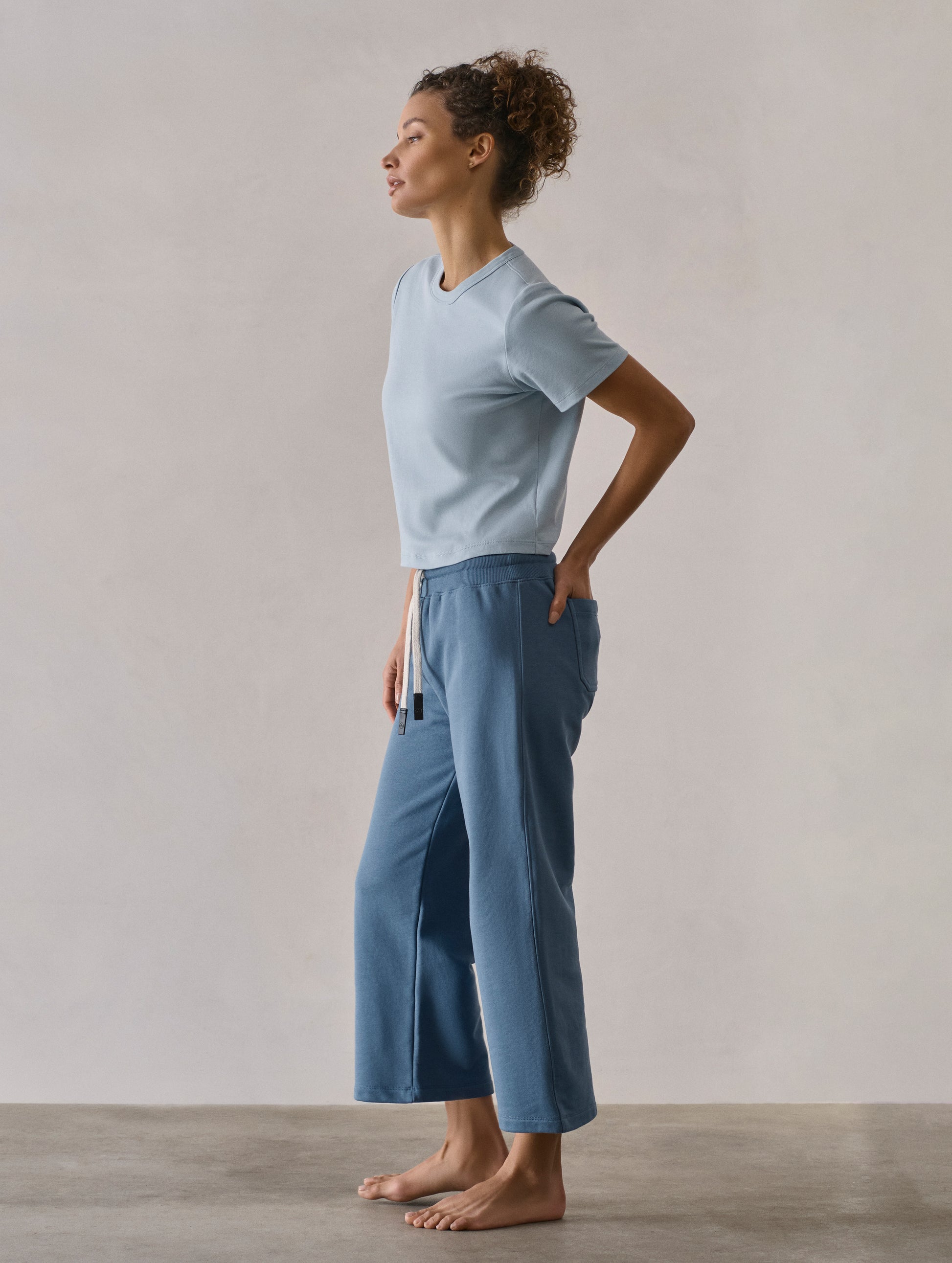 Left side profile full-body view of woman wearing W Osa Cropped Sweatpant in Coastal Blue from AETHER Apparel.