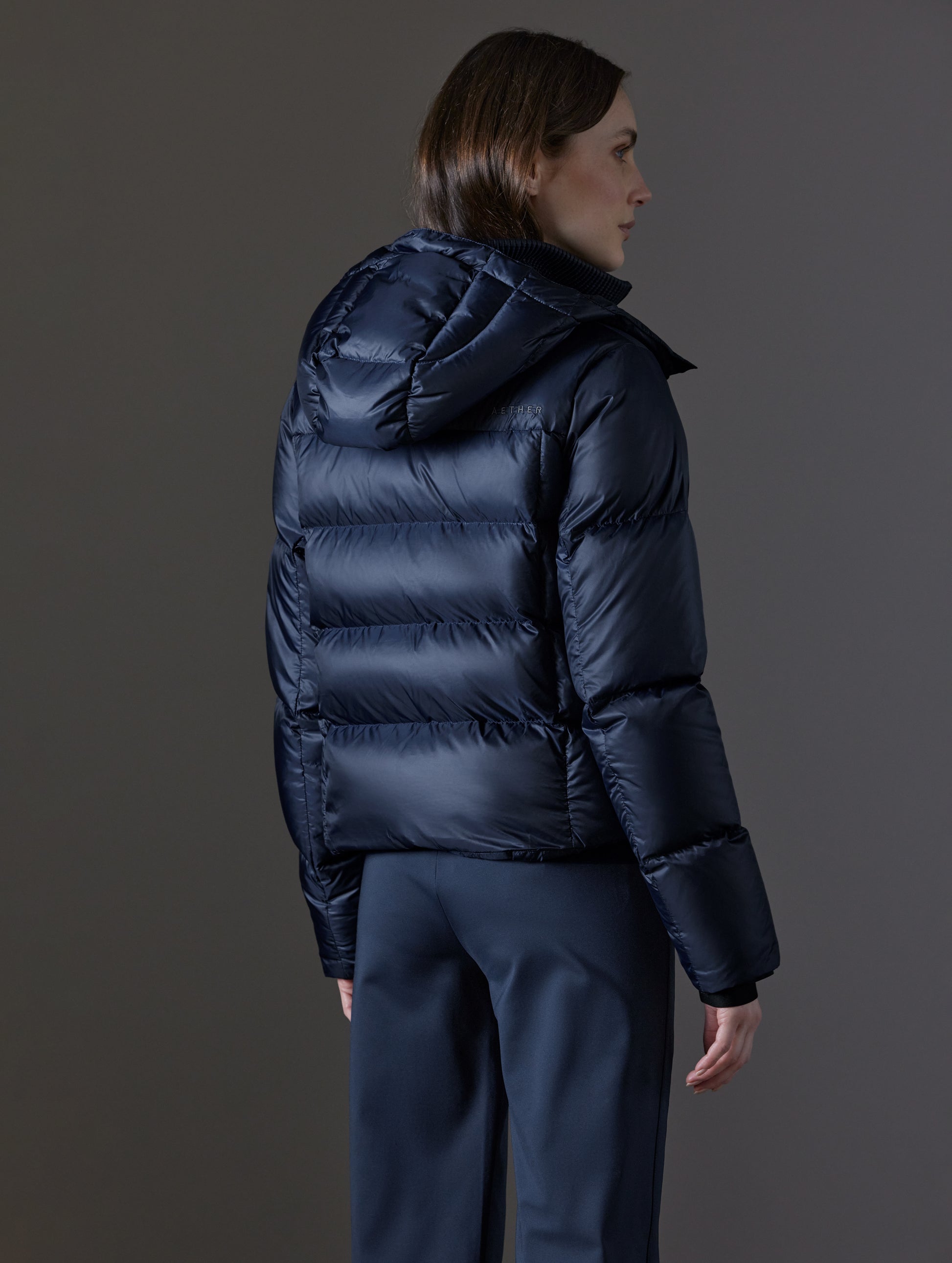 Rear angled view of woman wearing Aura Puffer Jacket in Total Eclipse dark blue from AETHER Apparel.