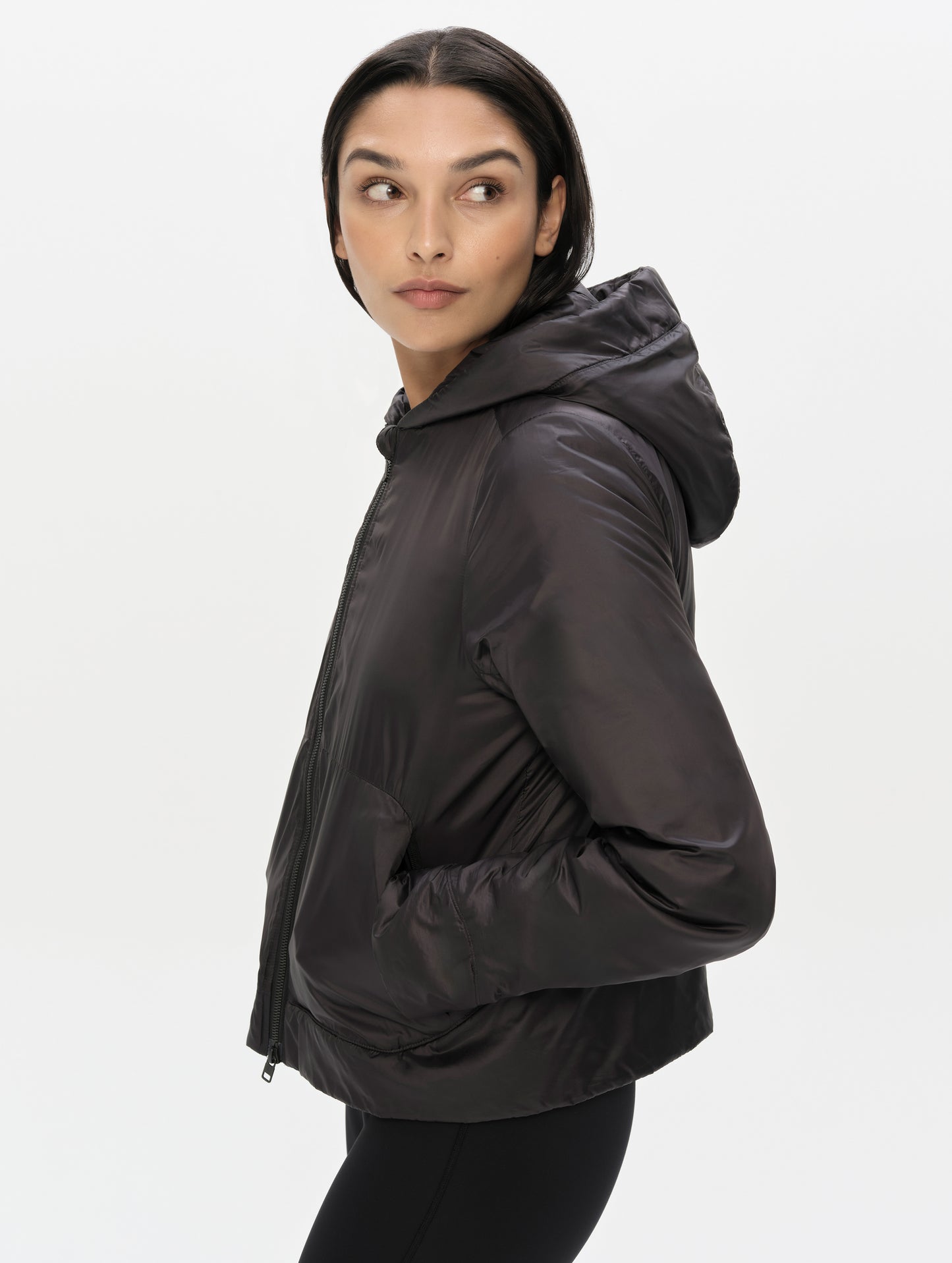 Side profile upper-body view of woman with hands in pockets wearing the Celeste Eco Insulated Hoodie in Onyx Black from AETHER Apparel.