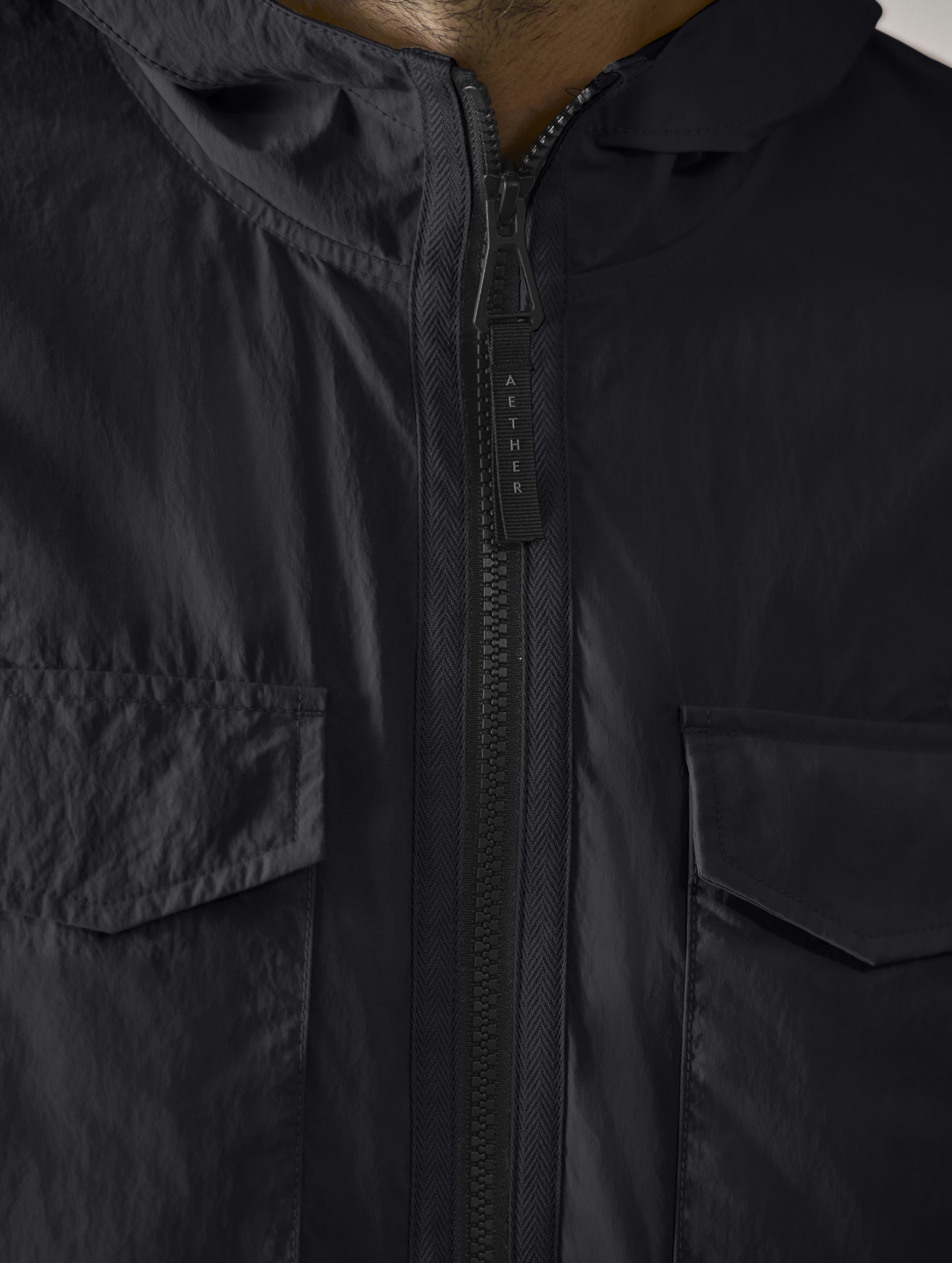 Closeup detailed view of the main center-front zipper and bellow patch chest pockets with hidden snap closures on the Aires Lightweight Windbreaker in Onyx Black from AETHER Apparel.