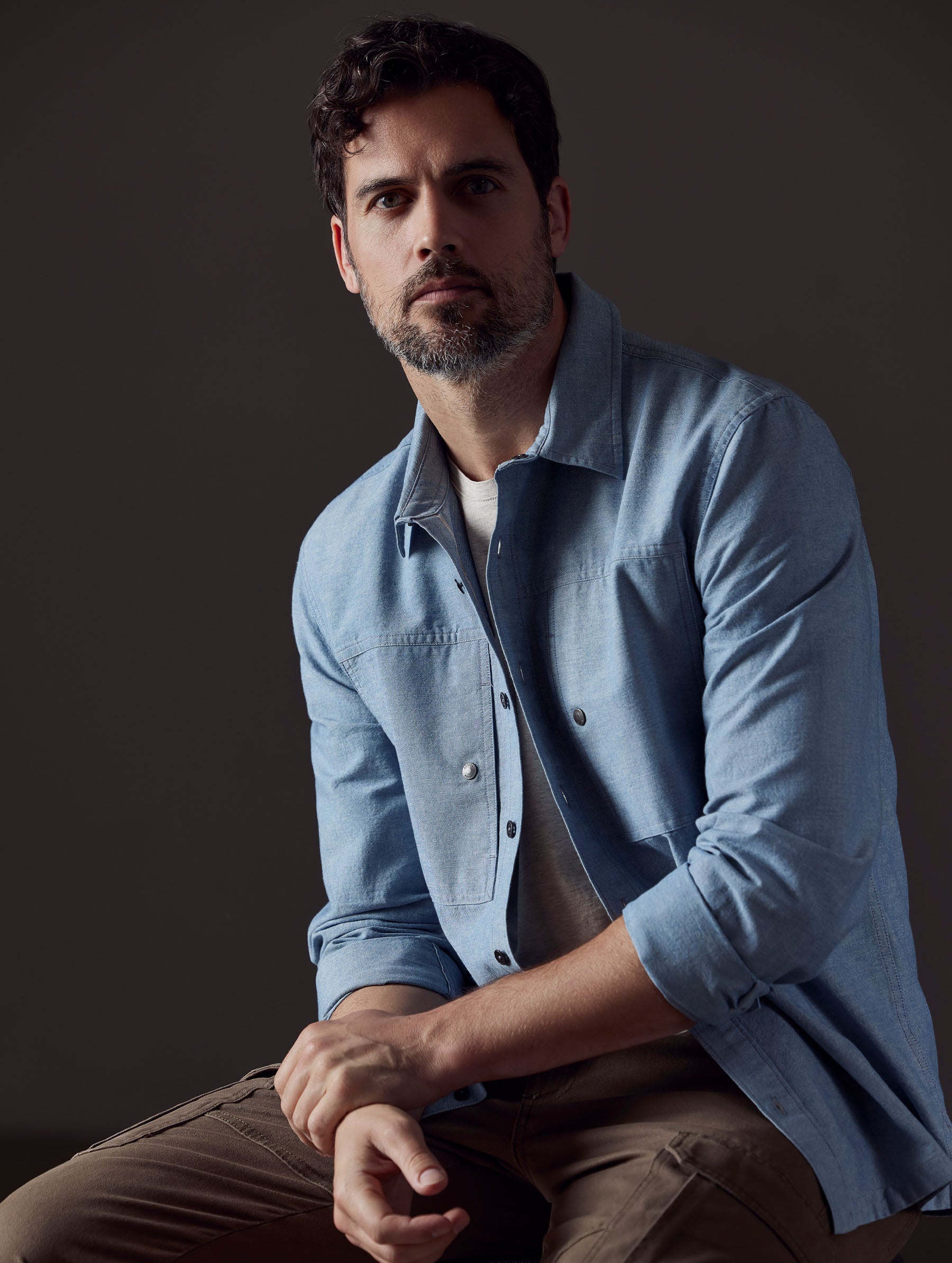 Man sitting with arms crossed wearing unbuttoned Emerson Button Down in Light Blue from AETHER Apparel.