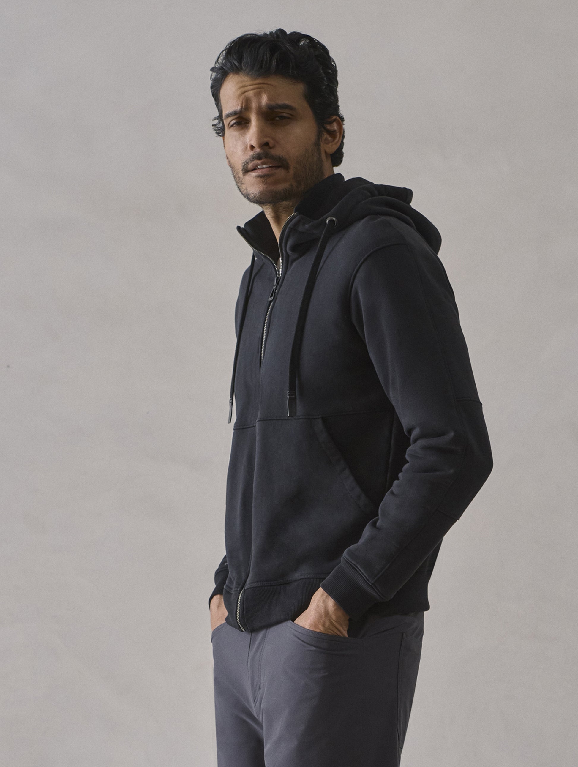 Front 3/4 upper-body view of man with hands in pant pockets wearing Monte Full-Zip Hoodie in Onyx Black from AETHER Apparel.