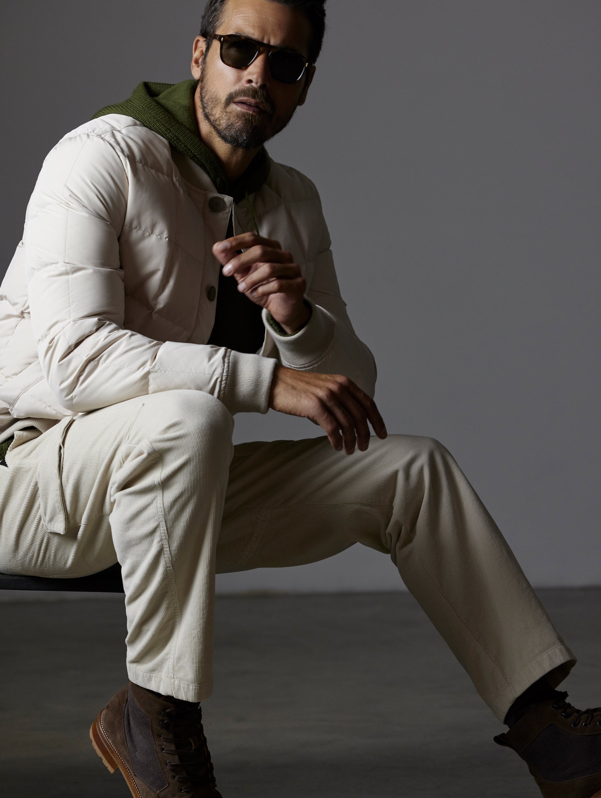 Front 3/4 full body view of man sitting wearing Pacer Jacket in Bone white from AETHER Apparel.