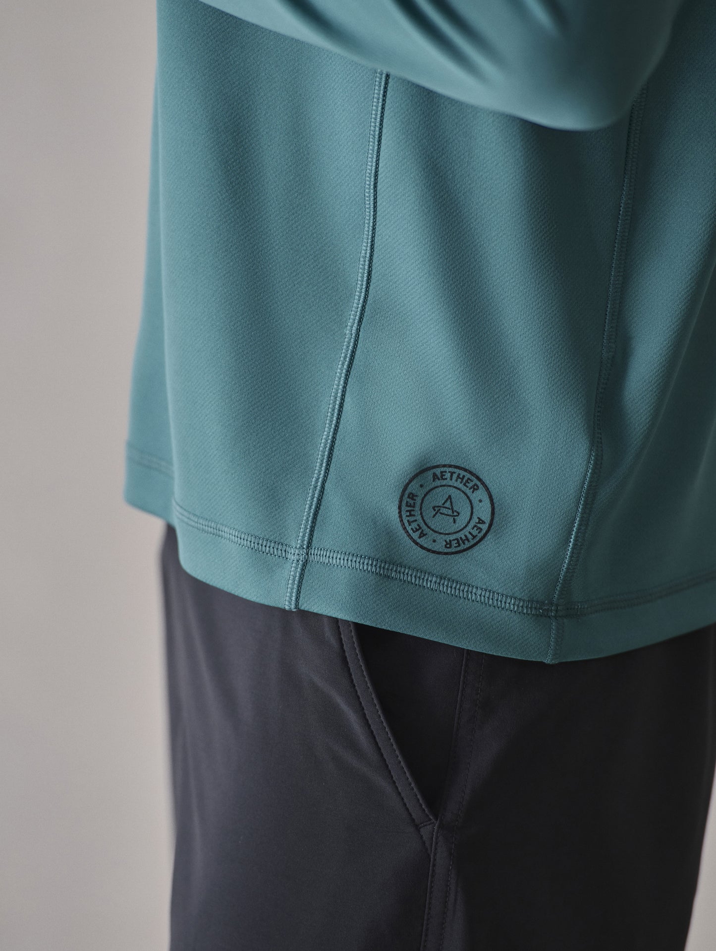 Closeup detailed view of the silicone heat transfer AETHER logo at the wearer’s left bottom side panel on the Pacific Crewneck Sun Shirt in Aqua Blue from AETHER Apparel.