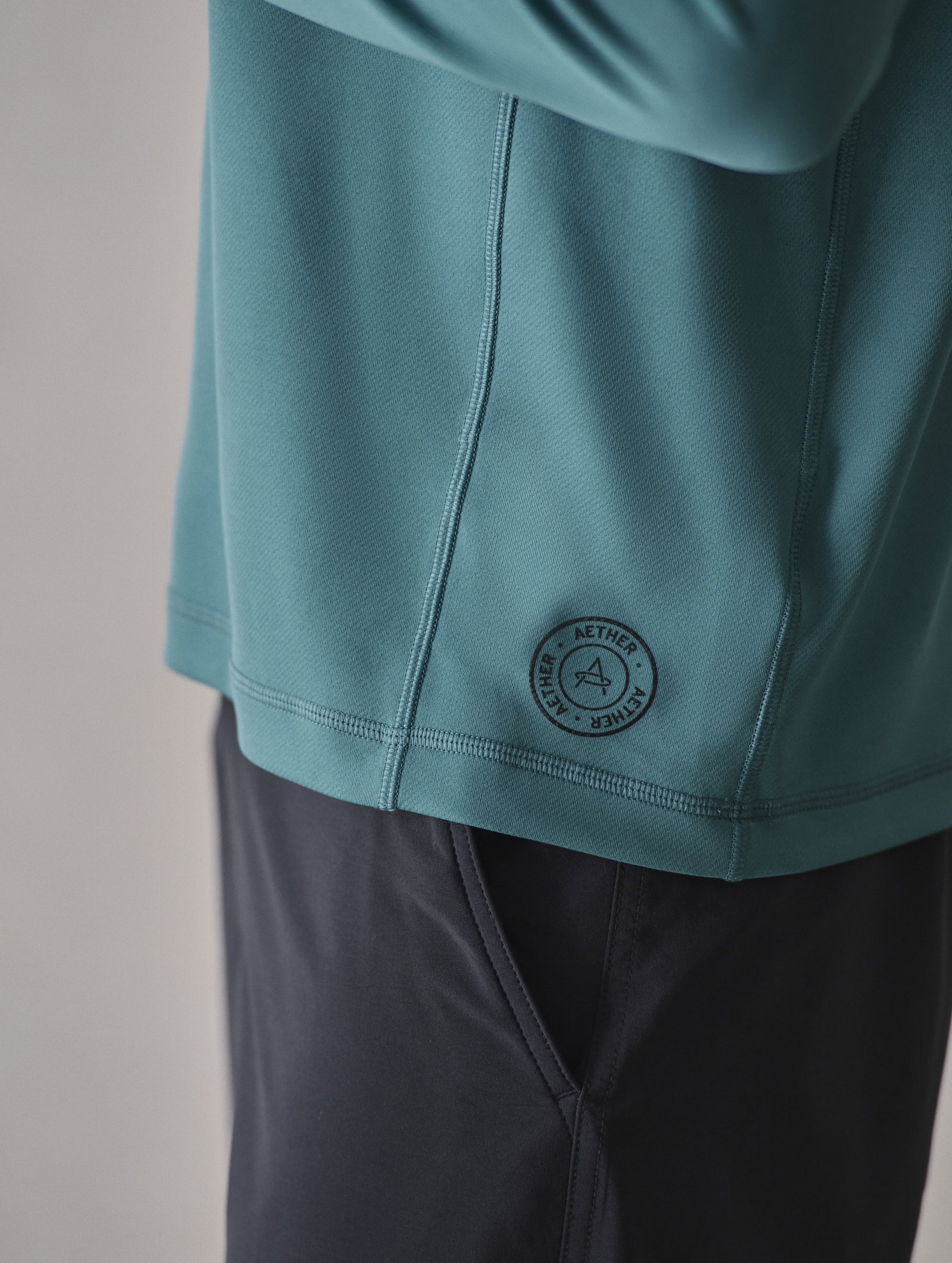 Closeup detailed view of the silicone heat transfer AETHER logo at the wearer’s left bottom side panel on the Pacific Crewneck Sun Shirt in Aqua Blue from AETHER Apparel.