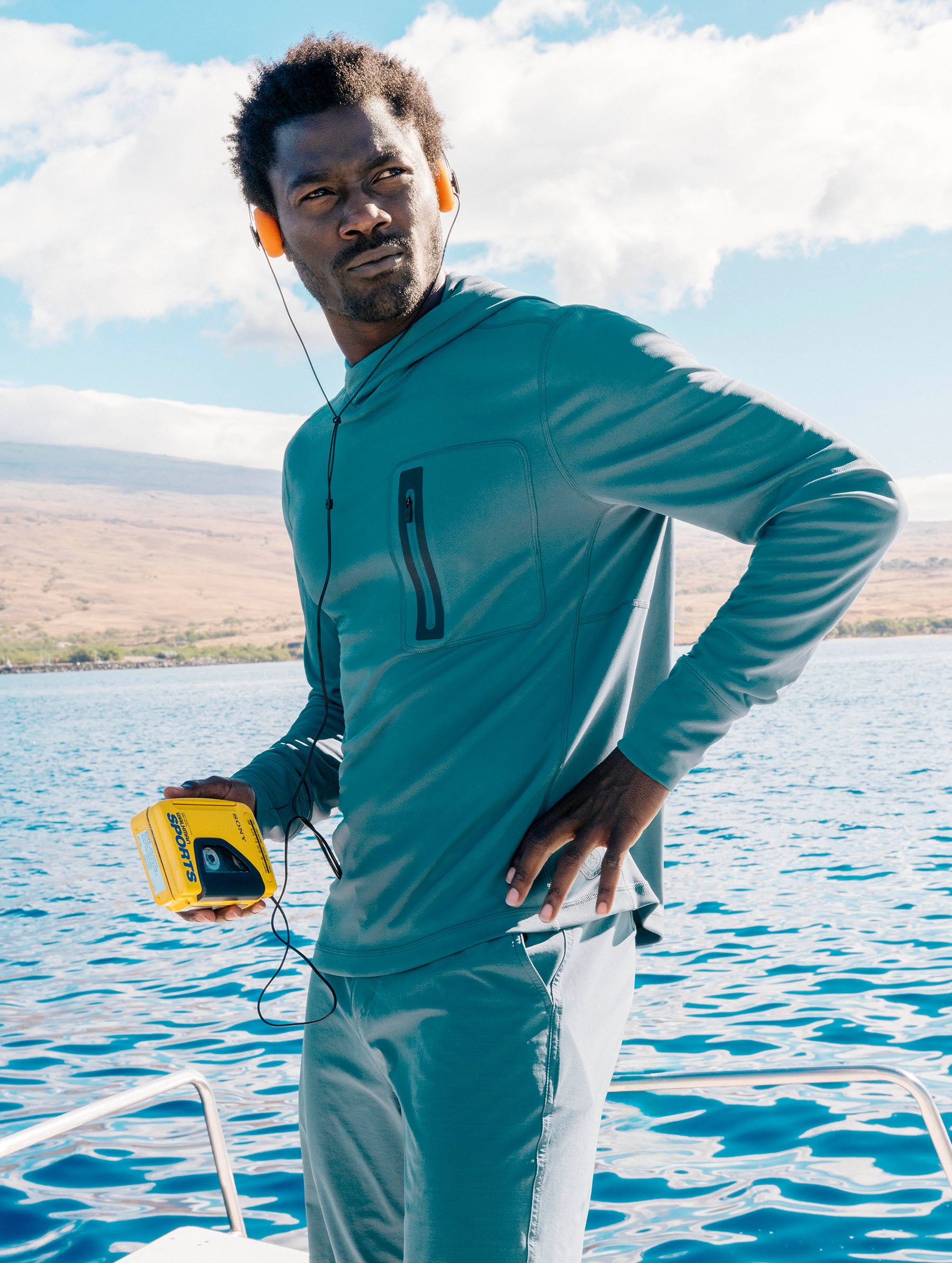 Lifestyle image of man standing on boat in the ocean with headphones on and Walkman in hand wearing the Pacific Hooded Sun Shirt in Aqua Blue from AETHER Apparel.