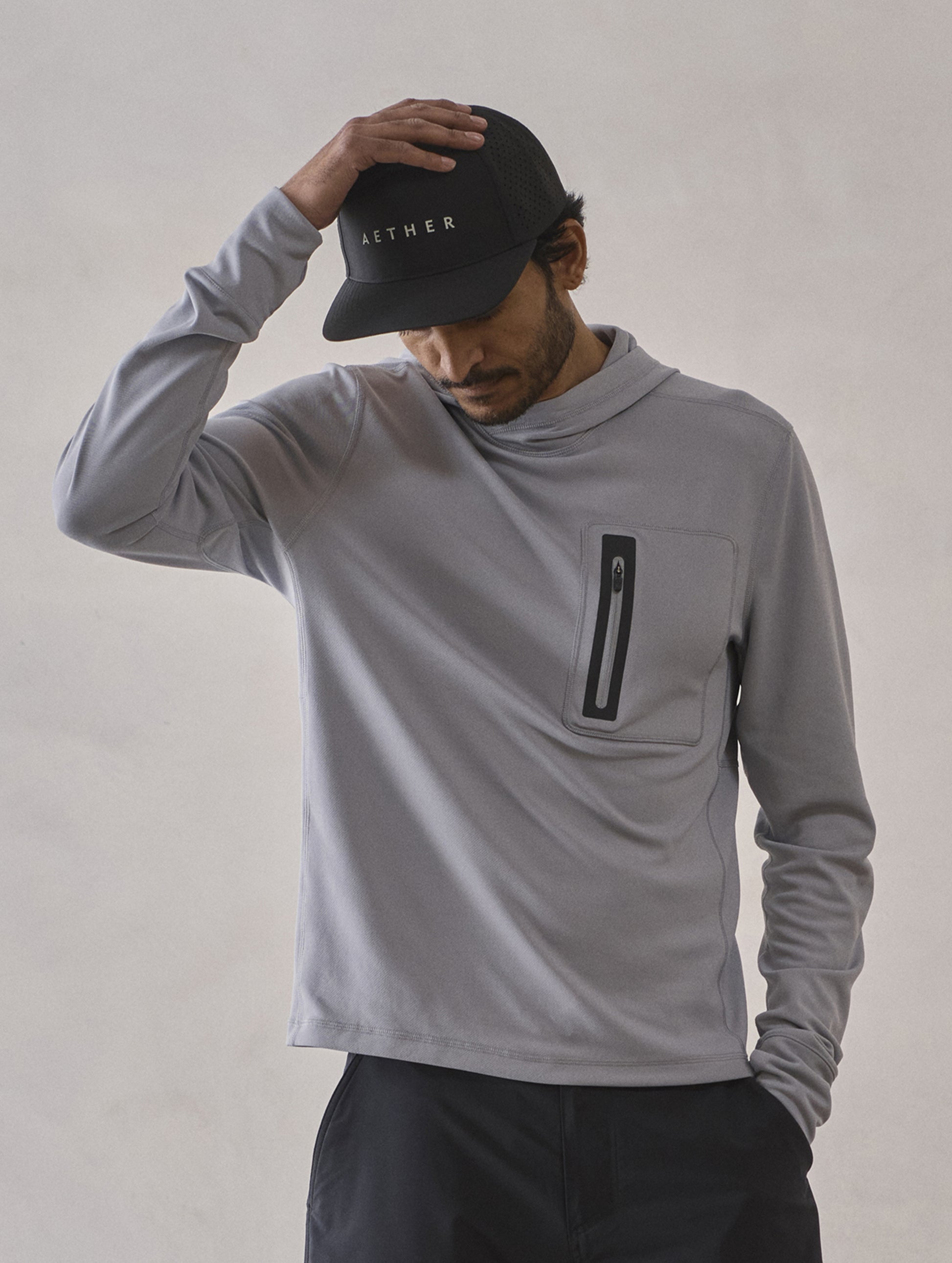 Front upper-body view of man with hand on hat wearing Pacific Hooded Sun Shirt in Sky Grey from AETHER Apparel.