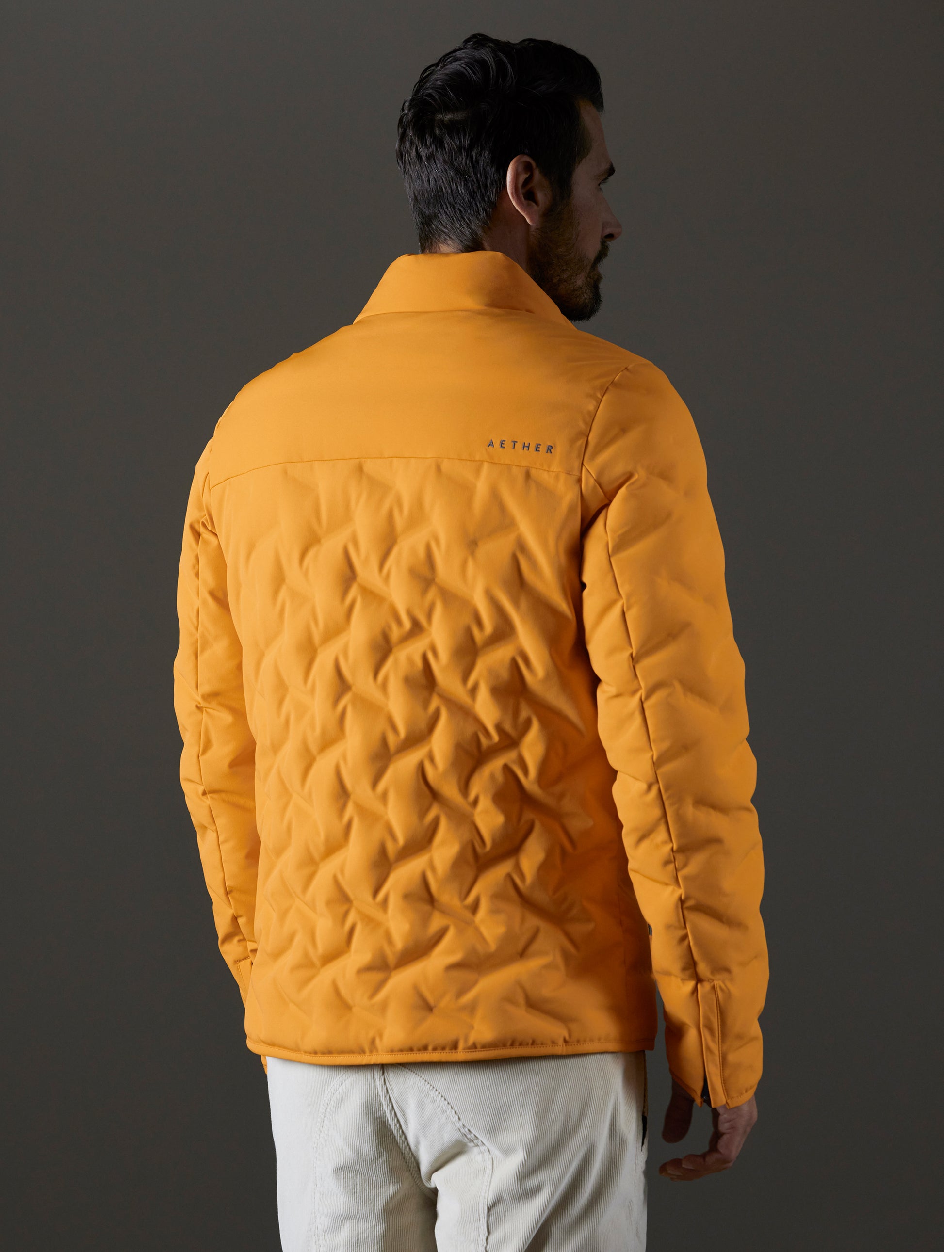 Rear 3/4 body view of man wearing Rex Down Jacket in Midas Orange from AETHER Apparel.