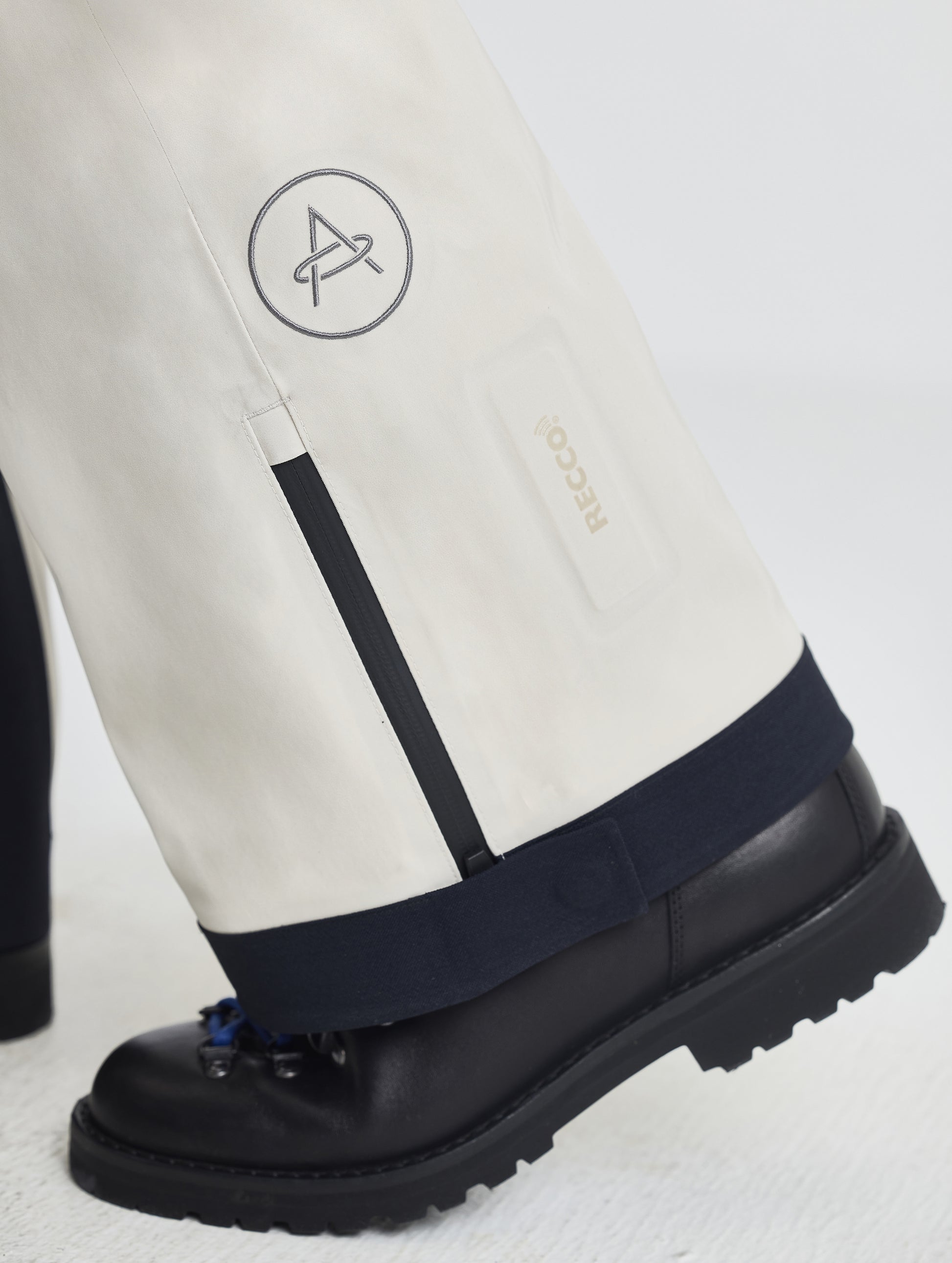 Closeup detailed view of the AETHER logo and zipper-adjustable leg cuffs on the Ridge Insulated Snow Pant in Bone from AETHER Apparel.