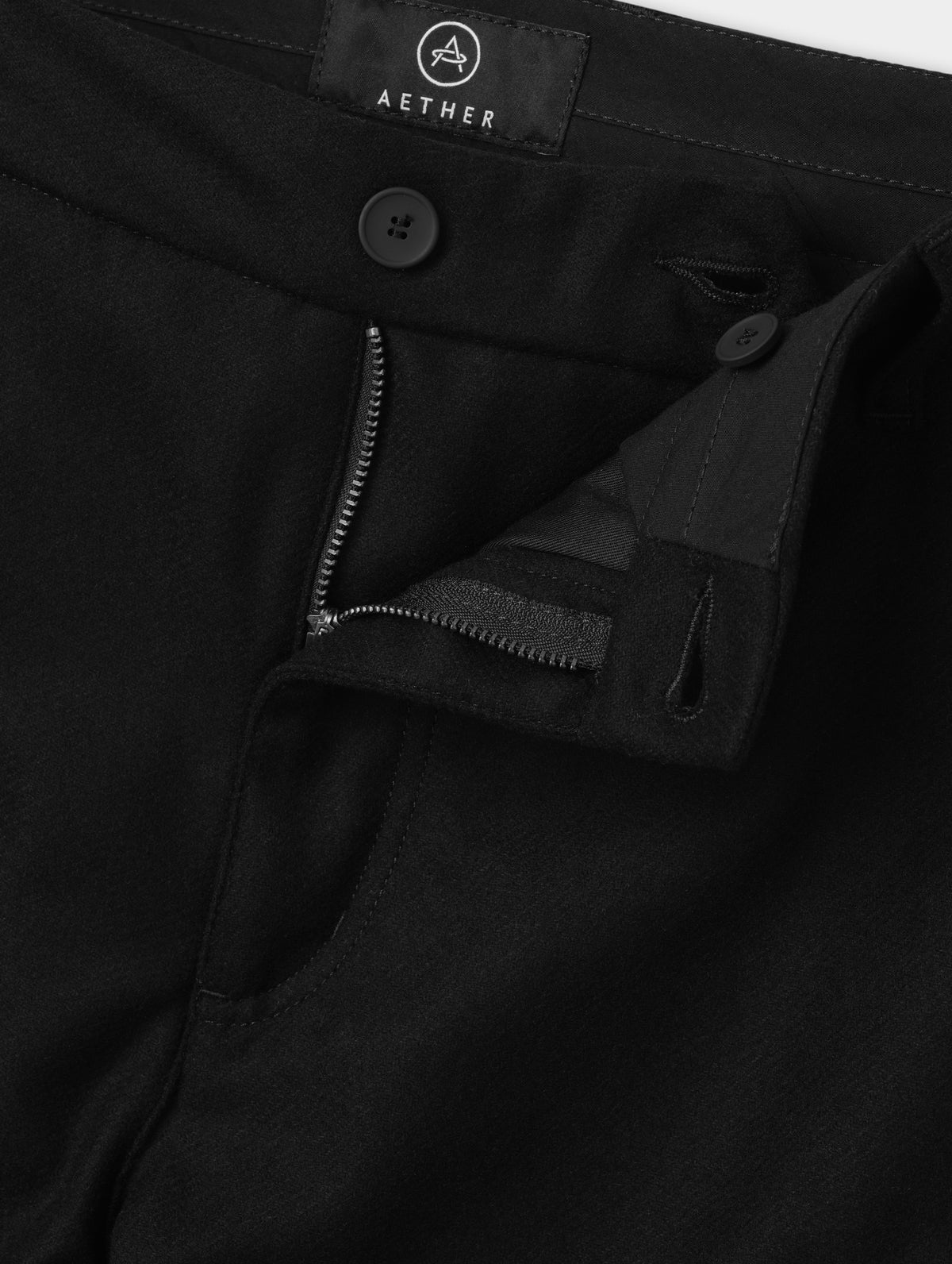 Closeup detailed view of the zippered fly, rubberized button, and waistband extension with button closure on the Strata Wool Pant in Onyx Black from AETHER Apparel.