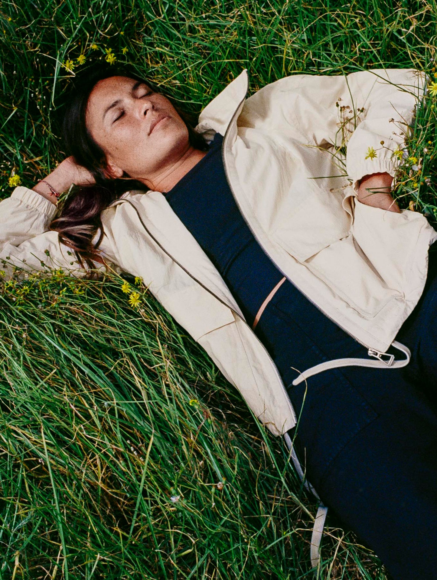 Lifestyle image of woman laying in tall grass field wearing the Mila Windbreaker in Bone white from AETHER Apparel.