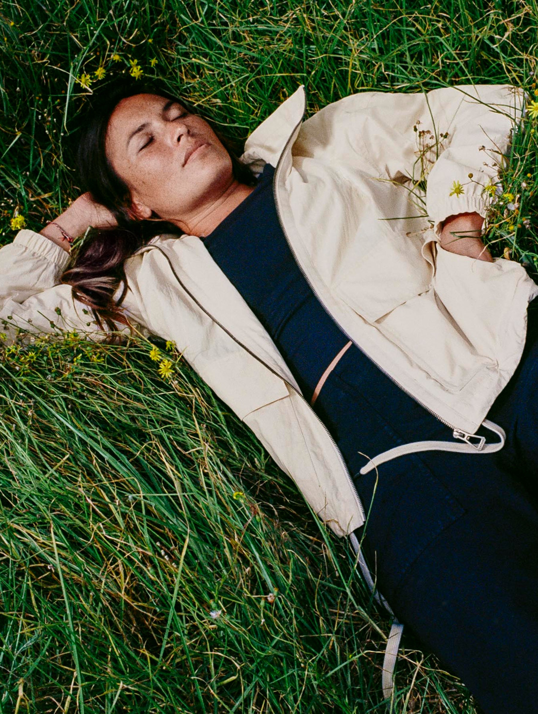 Lifestyle image of woman laying in tall grass field wearing the Mila Windbreaker in Bone white from AETHER Apparel.