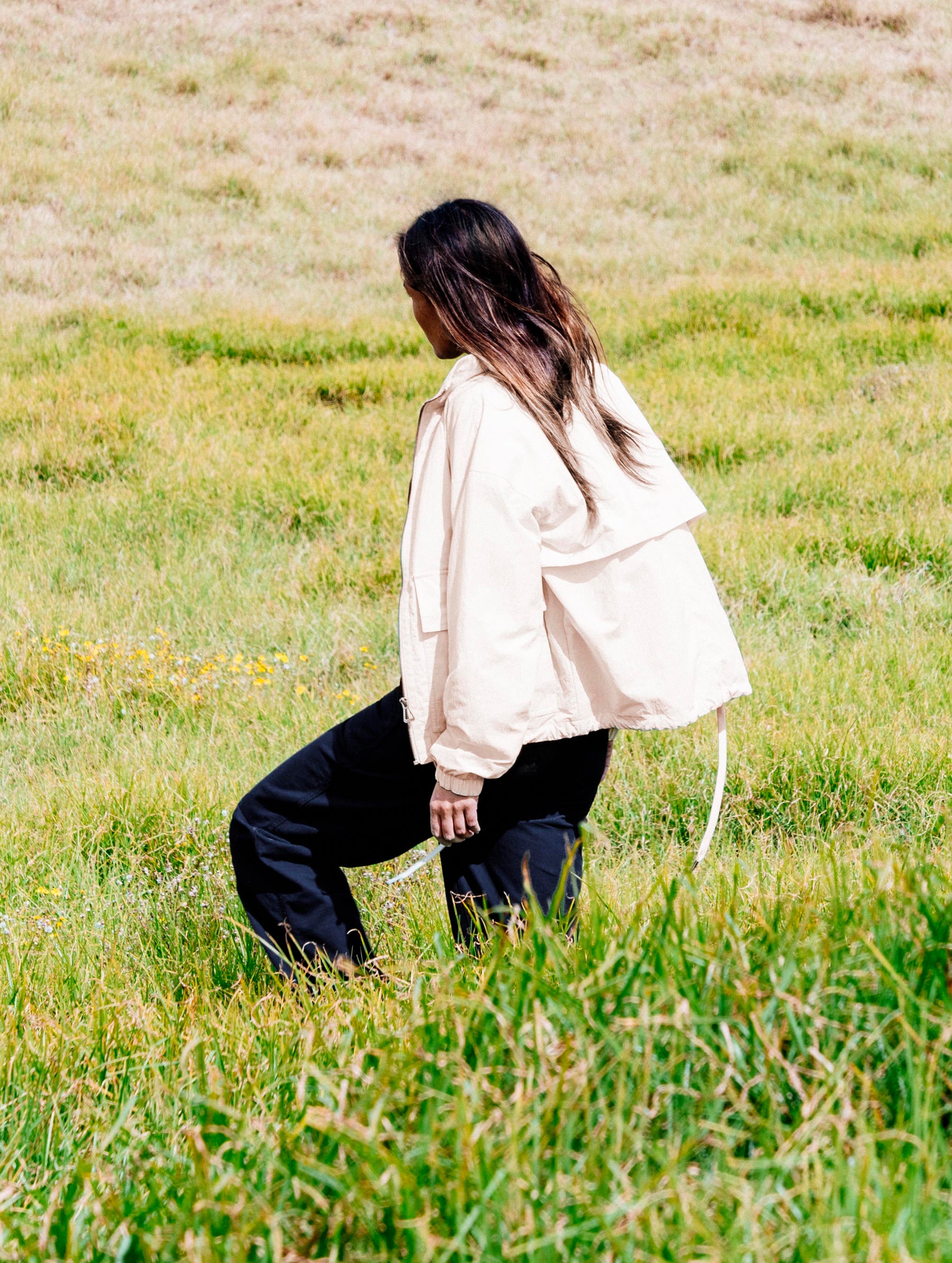 Lifestyle image of woman walking in tall grass field wearing the Mila Windbreaker in Bone white from AETHER Apparel.