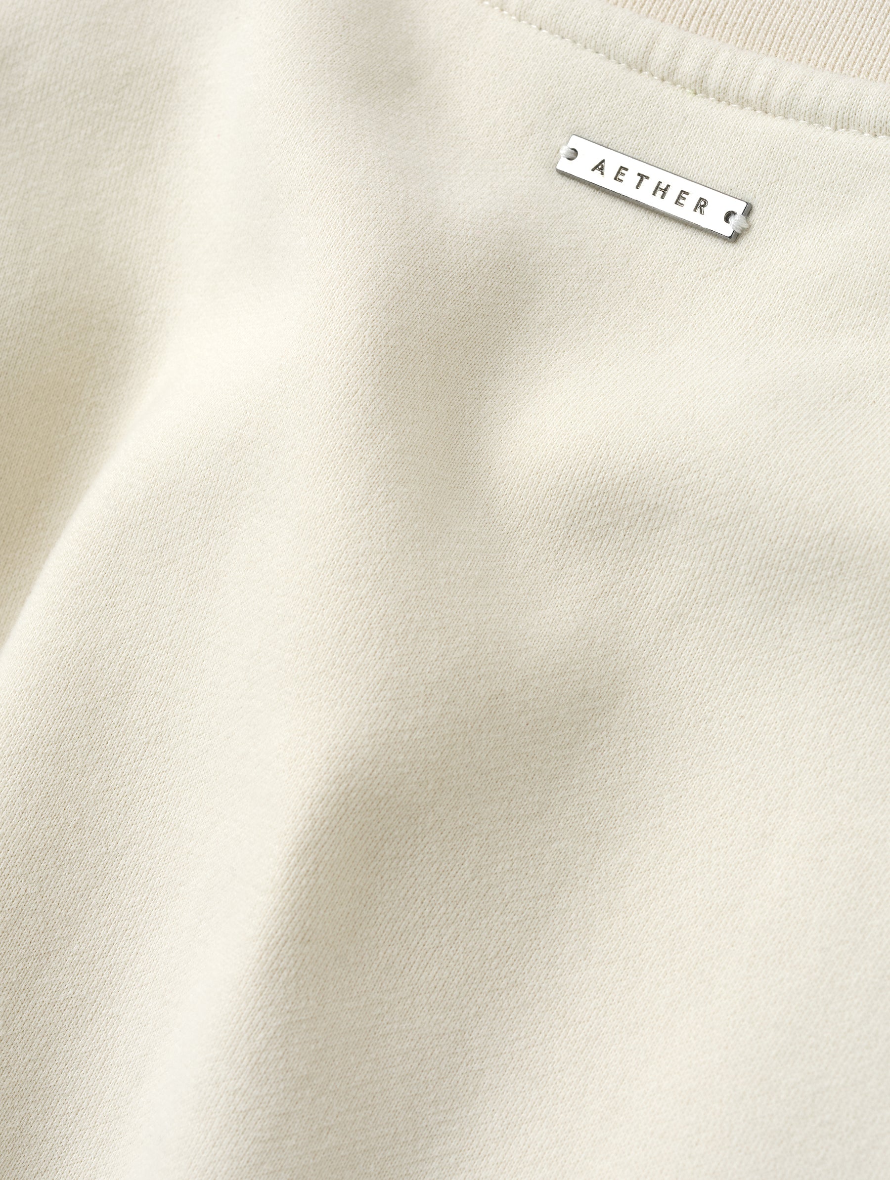 Closeup detailed view of the metal logo plate on the Rylan Crewneck Sweatshirt in Bone from AETHER Apparel.