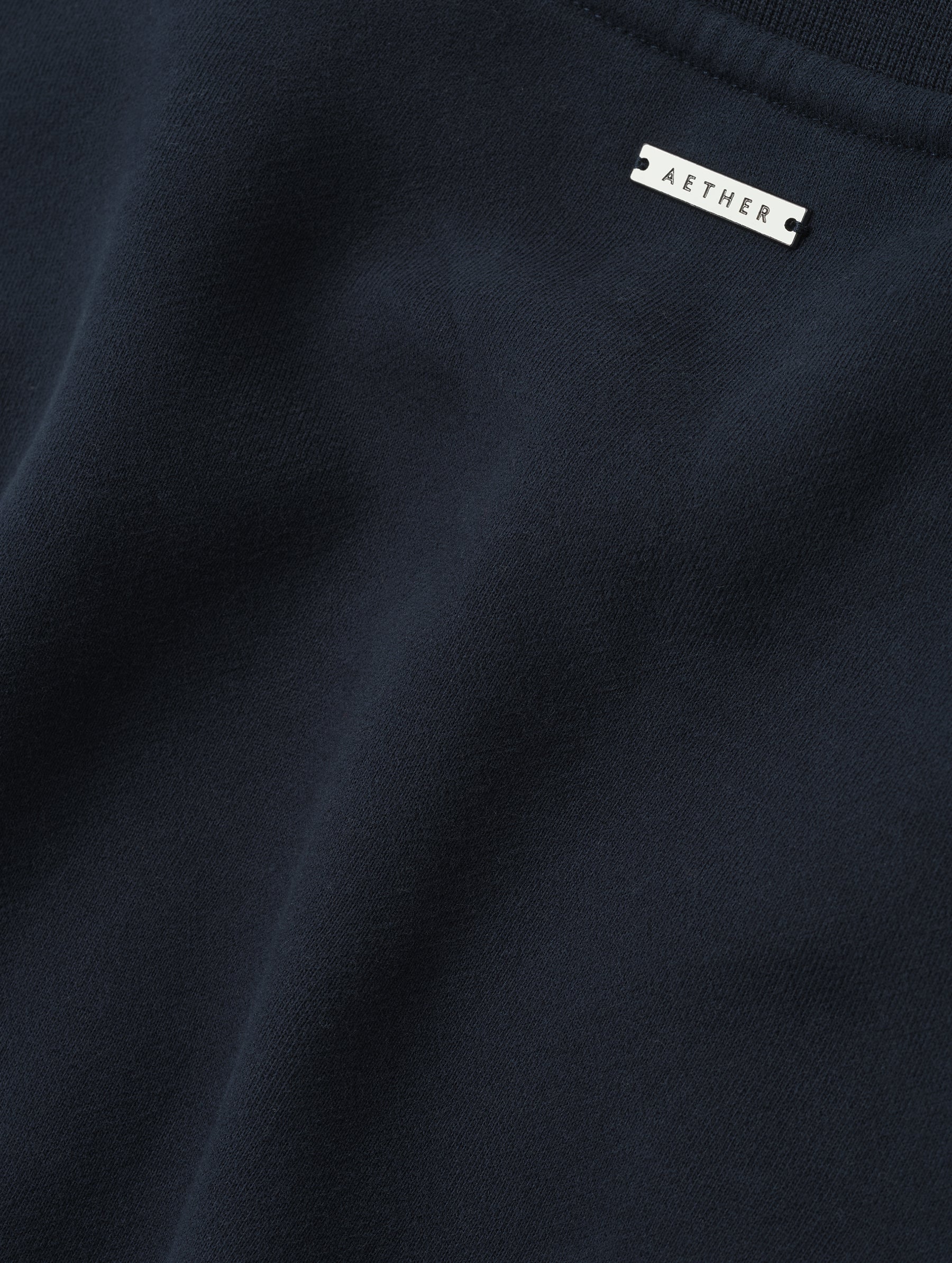 Closeup detailed view of the metal logo plate on the Rylan Crewneck Sweatshirt in Total Eclipse from AETHER Apparel.