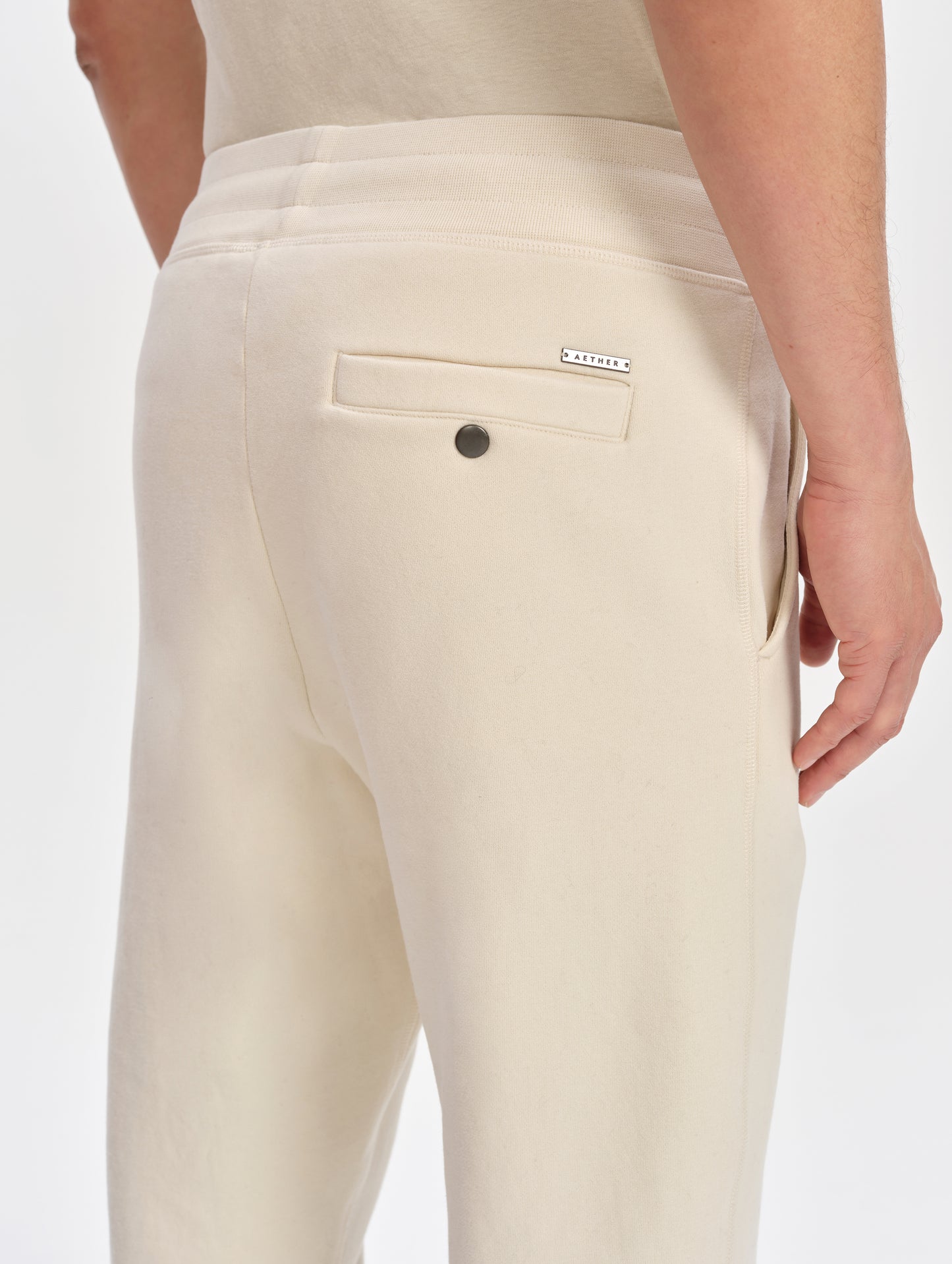 Closeup detailed view of the custom metal AETHER logo plate and snap-secured back pocket on the Rylan Sweat Pant in Bone from AETHER Apparel. 