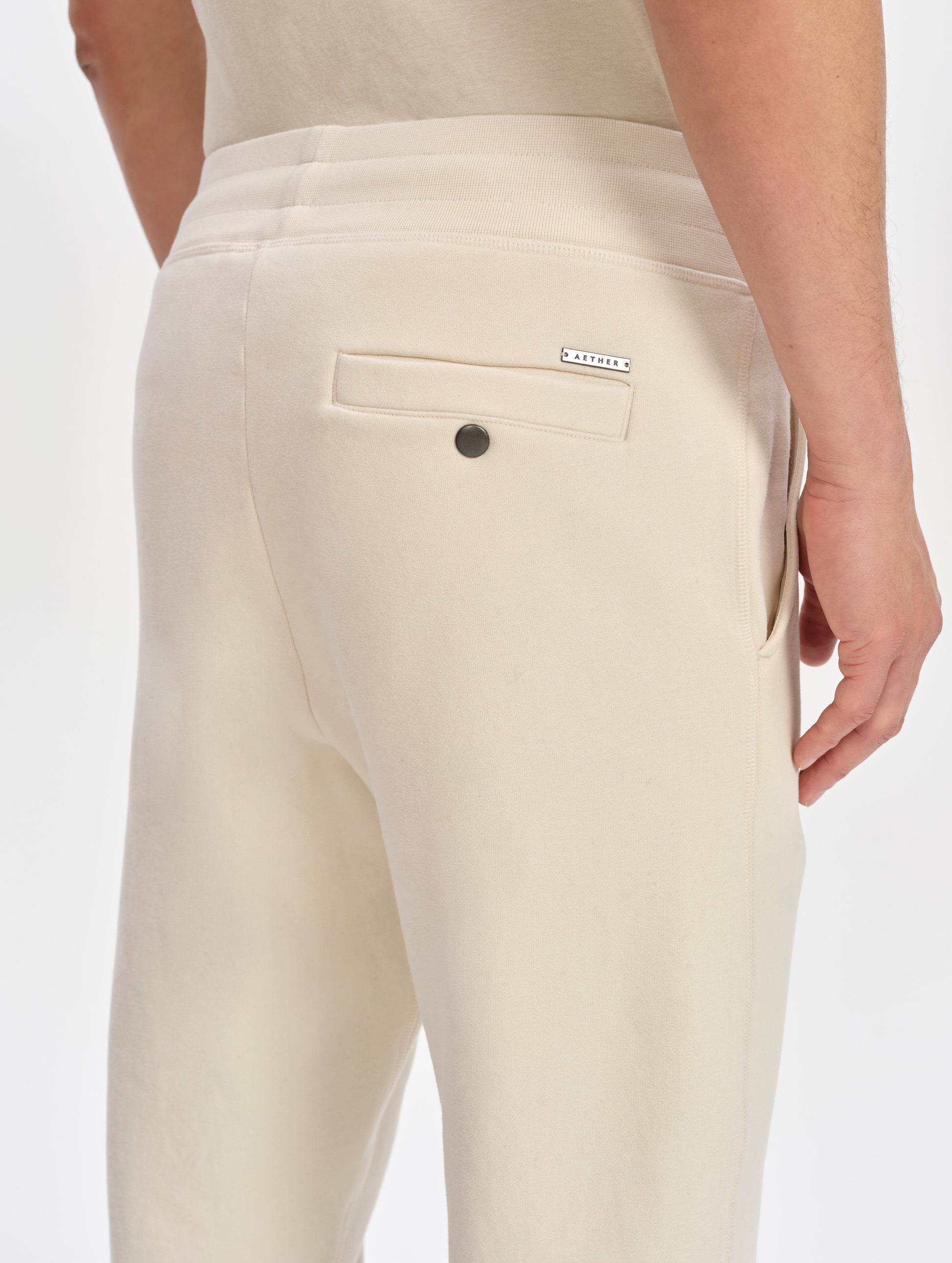 Closeup detailed view of the custom metal AETHER logo plate and snap-secured back pocket on the Rylan Sweat Pant in Bone from AETHER Apparel. 