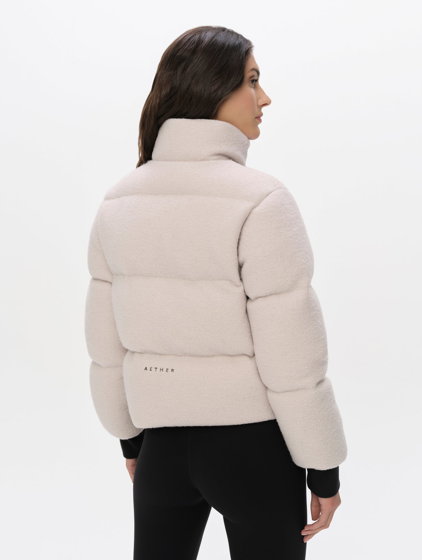 Rear angled upper-body view of woman wearing the Aura Puffer Jacket Wool in Bone white from AETHER Apparel.