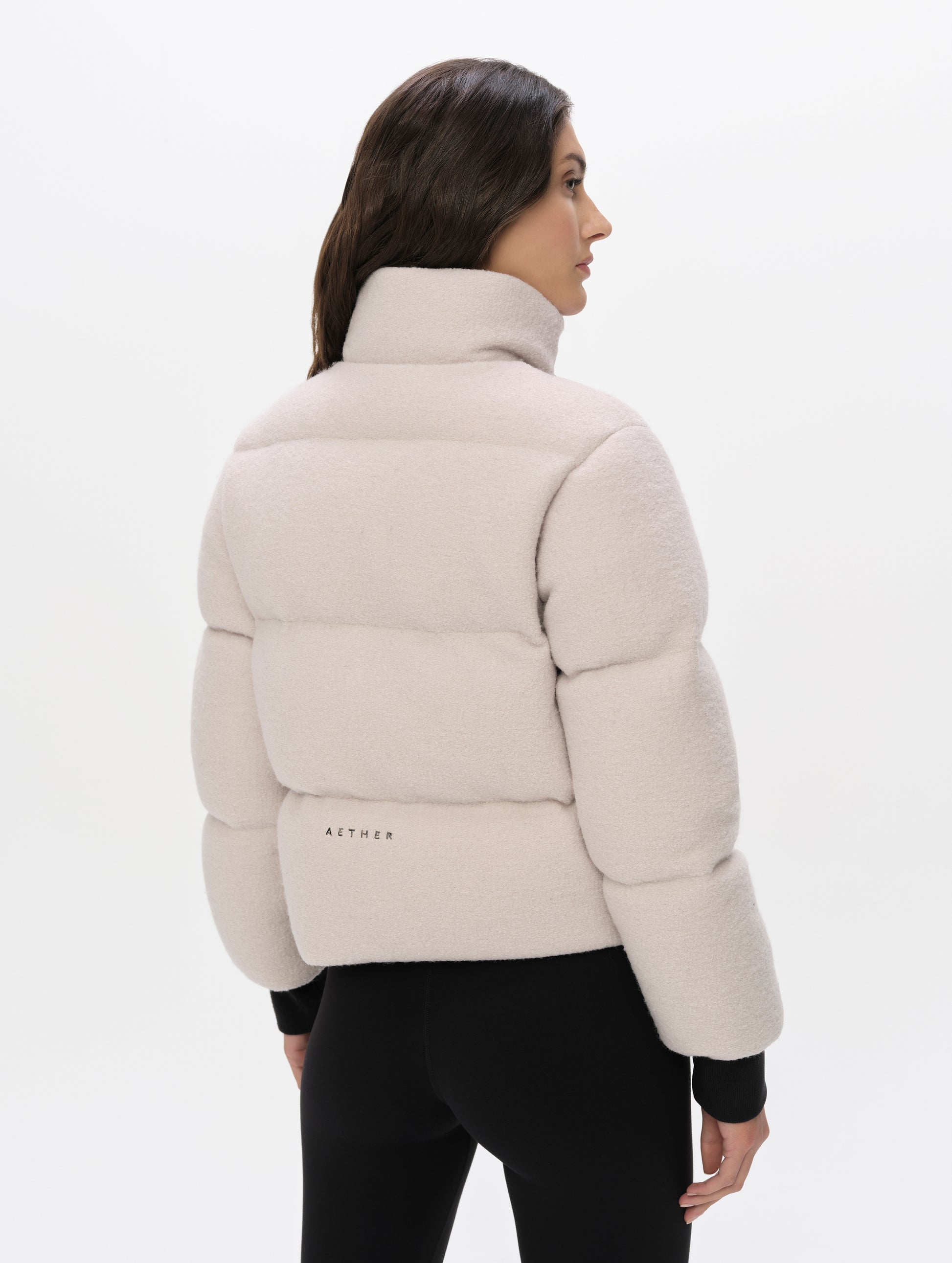 Rear angled upper-body view of woman wearing the Aura Puffer Jacket Wool in Bone white from AETHER Apparel.