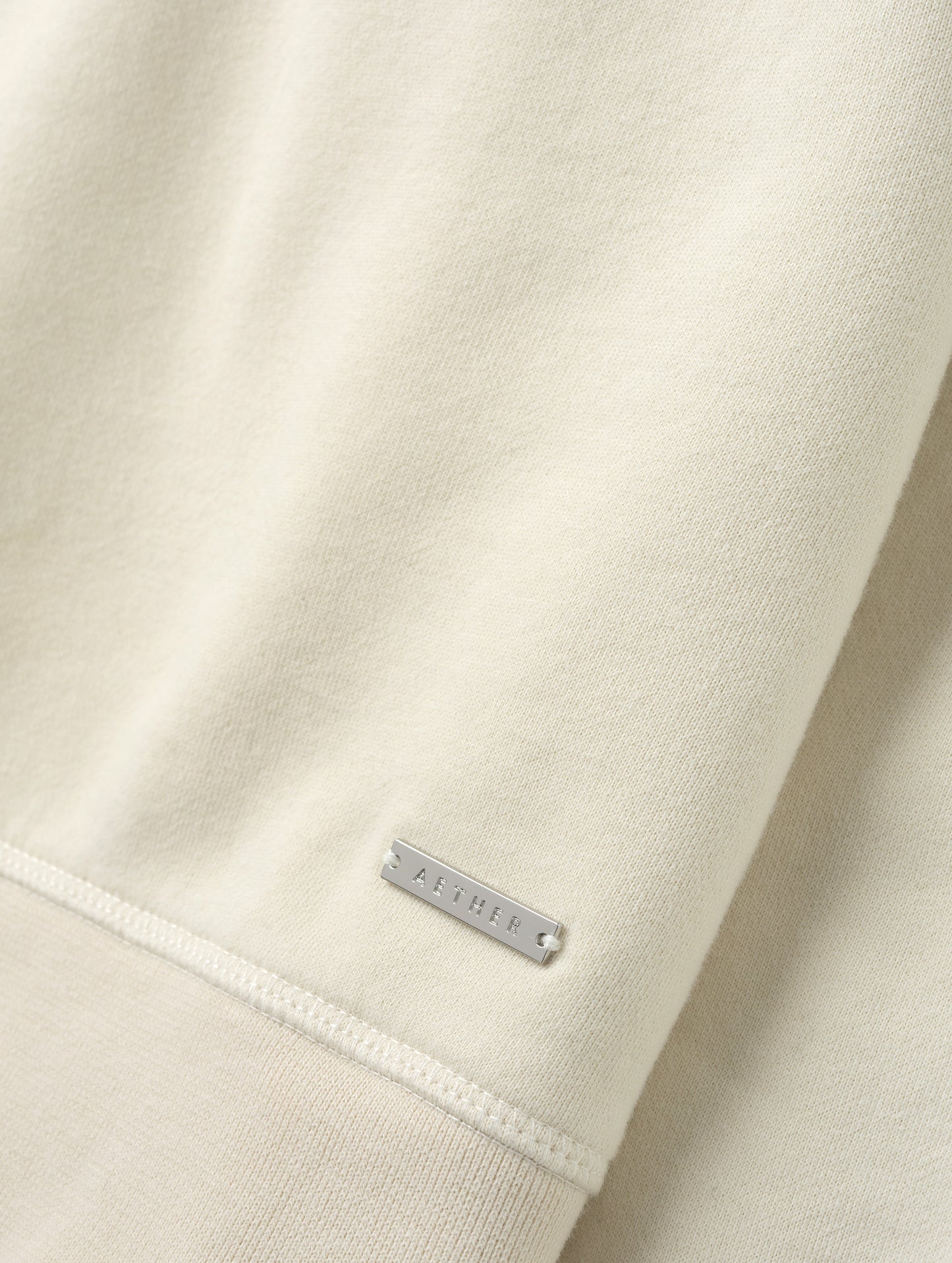 Closeup detailed view of the metal logo plate on the Ayla Crewneck Sweatshirt in Bone from AETHER Apparel.