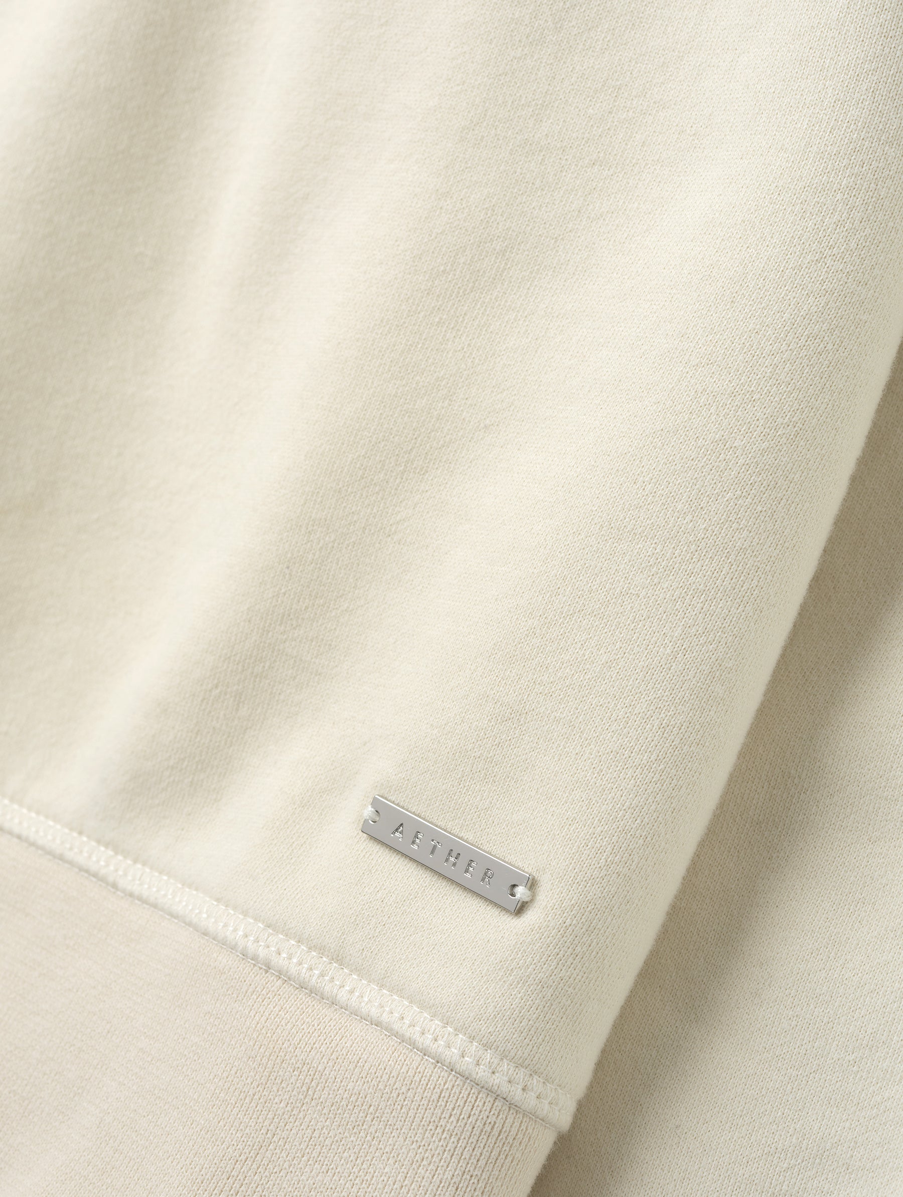 Closeup detailed view of the metal logo plate on the Ayla Crewneck Sweatshirt in Bone from AETHER Apparel.