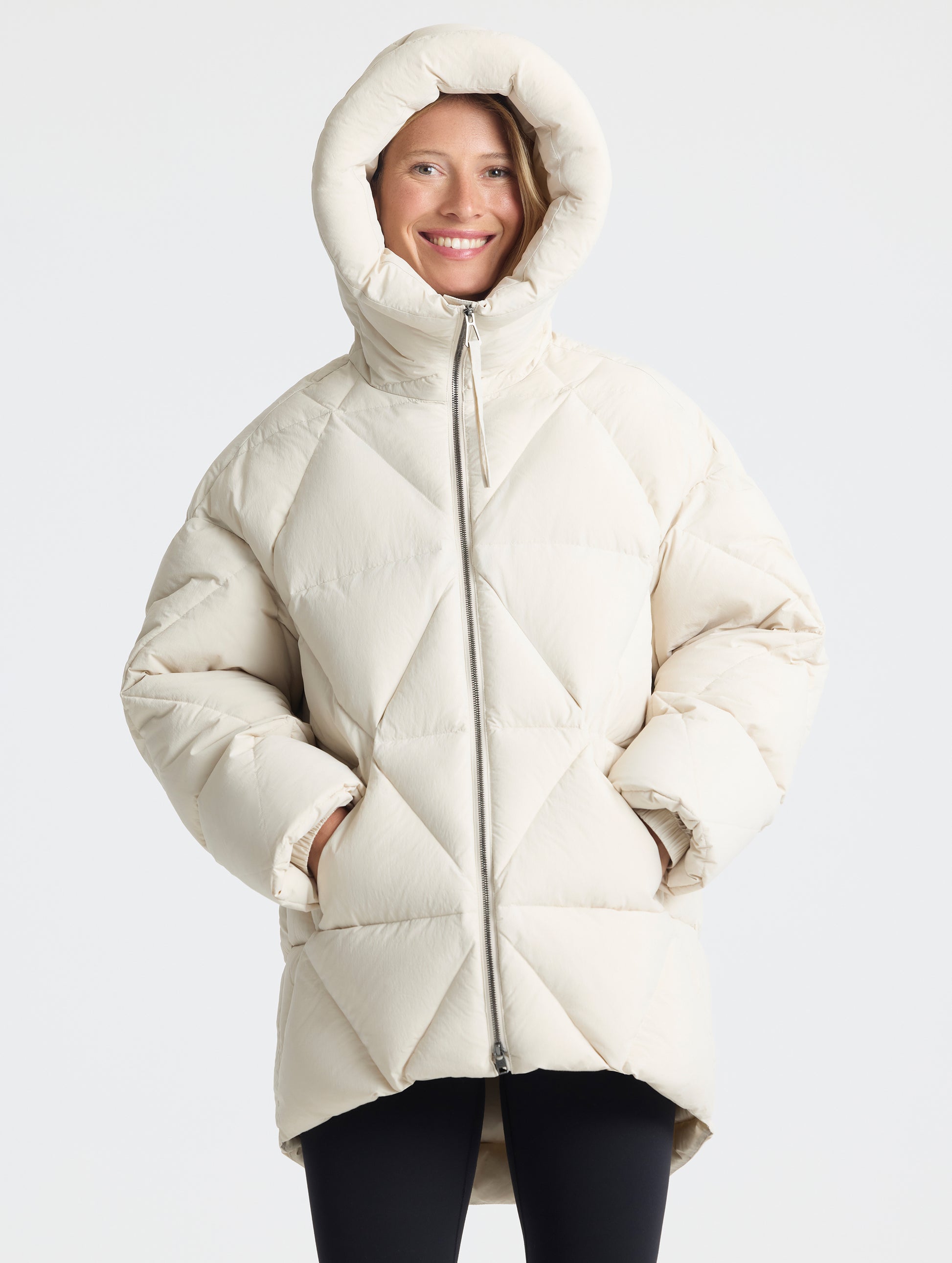 Front upper-body view of woman with hood up wearing a fully zipped up Callie Oversized Down Parka in Bone from AETHER Apparel.