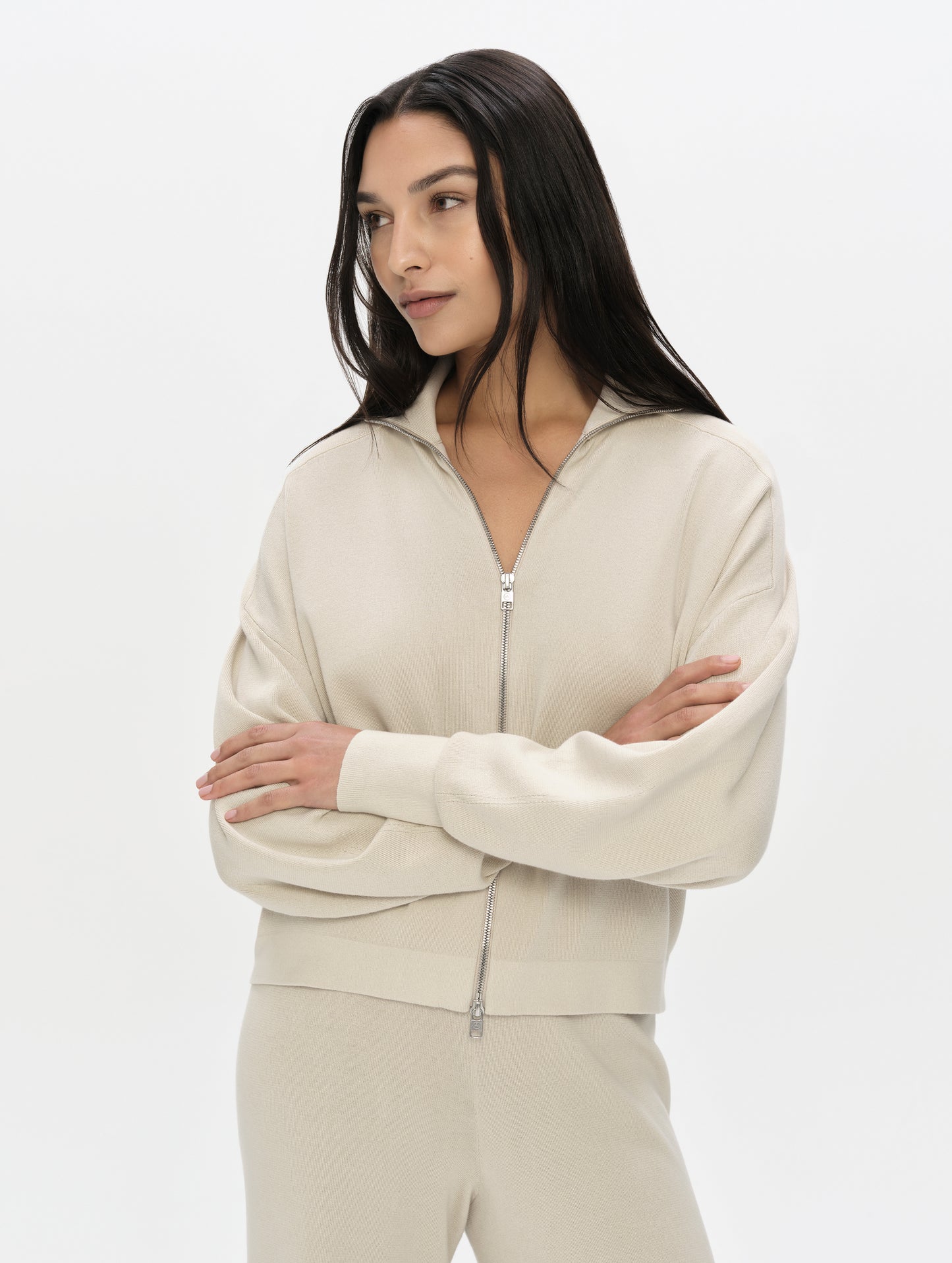 Front, upper-body view of woman with arms crossed wearing the Eliana Full-ZIp Sweater in Bone from AETHER Apparel.