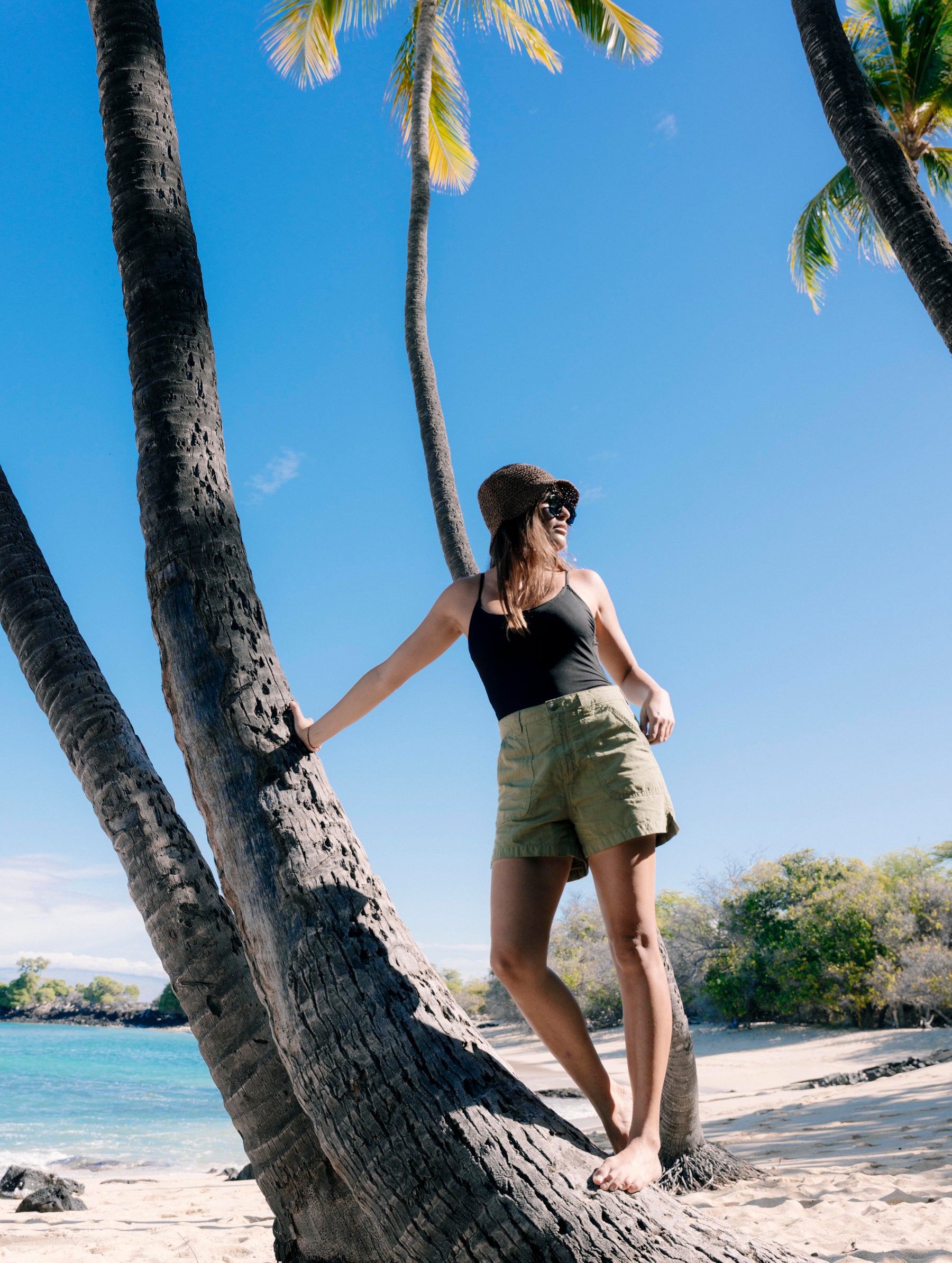 Lifestyle image of woman standing and leaning against a palm tree while wearing the Masai Safari Short in Fern Green from AETHER Apparel.