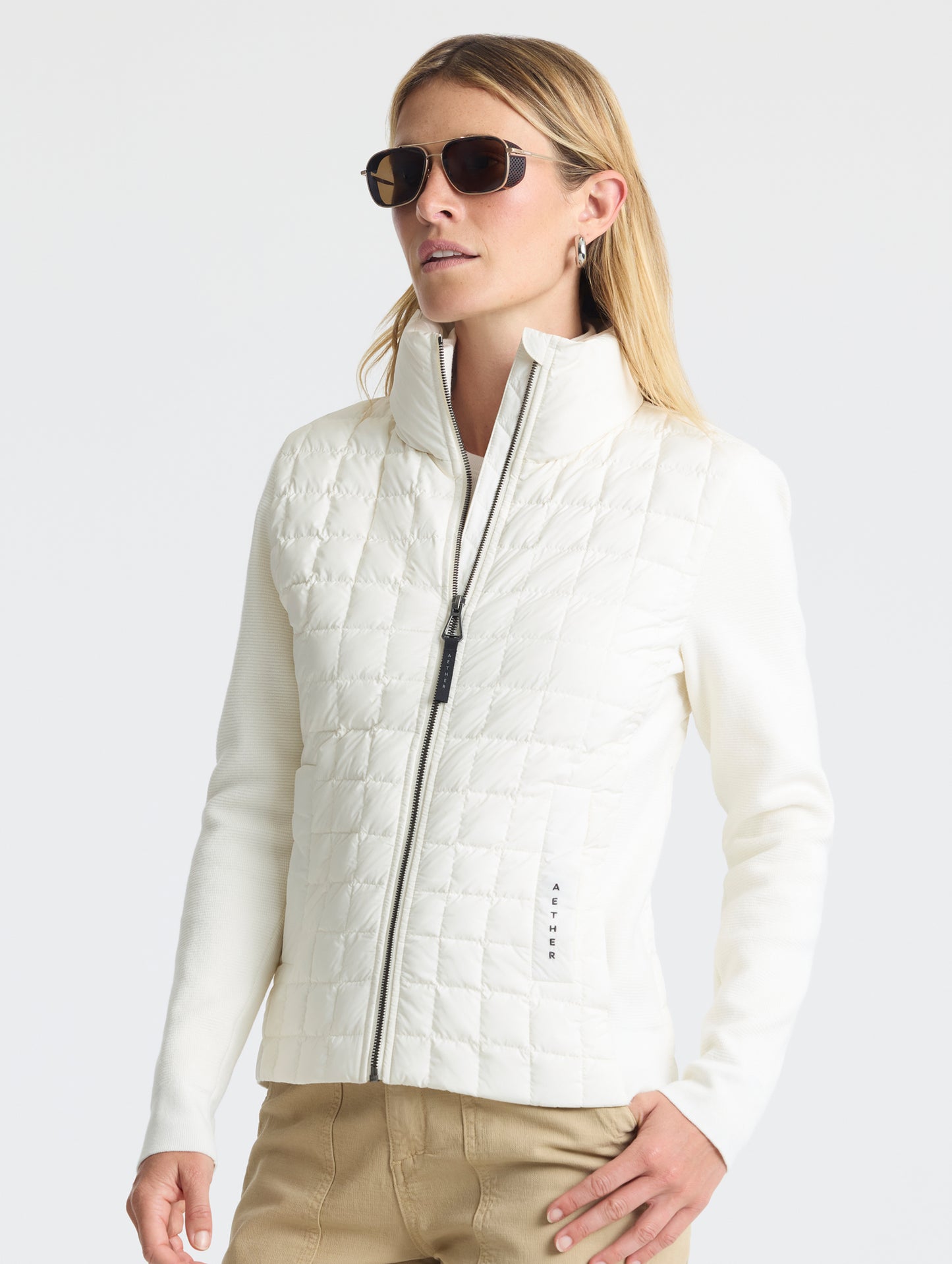 Front, angled, upper-body view of woman sunglasses and partially-unzipped Phase Full-Zip in Winter White from AETHER Apparel.