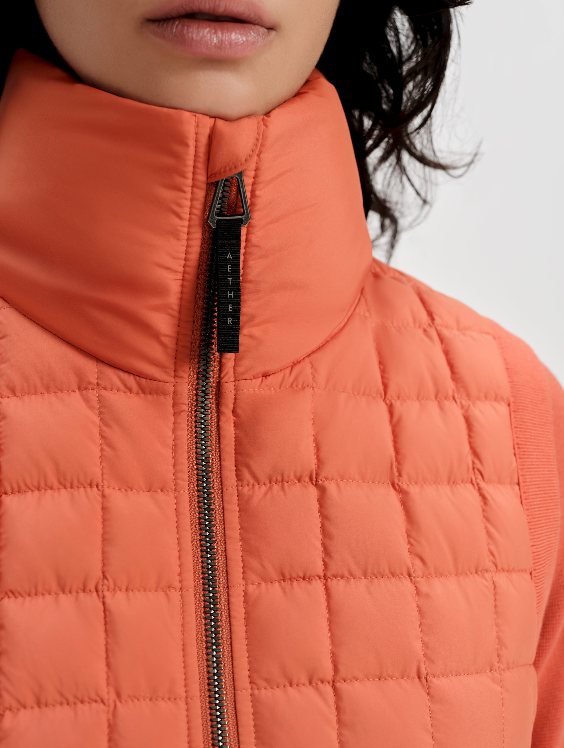 Closeup detailed view of the insulated collar and AETHER-branded zipper pull on the center-front of the Phase Vest in Madeira Orange from AETHER Apparel.