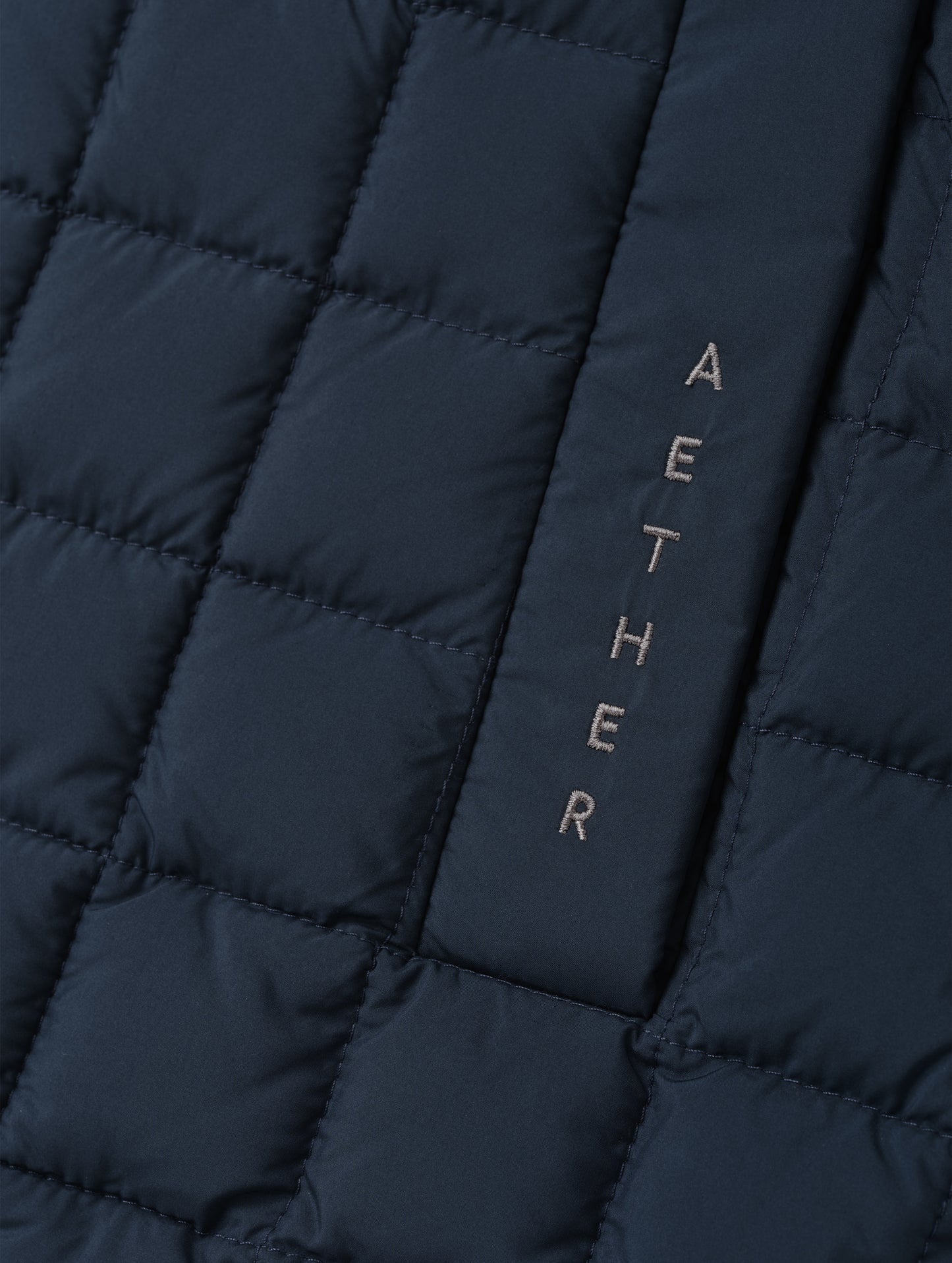 Closeup detailed view of the embroidered wordmark and box quilting on the Phase Vest in Total Eclipse from AETHER Apparel.