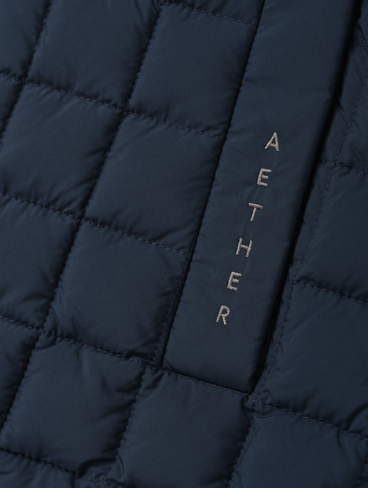 Closeup detailed view of the embroidered wordmark and box quilting on the Phase Vest in Total Eclipse from AETHER Apparel.