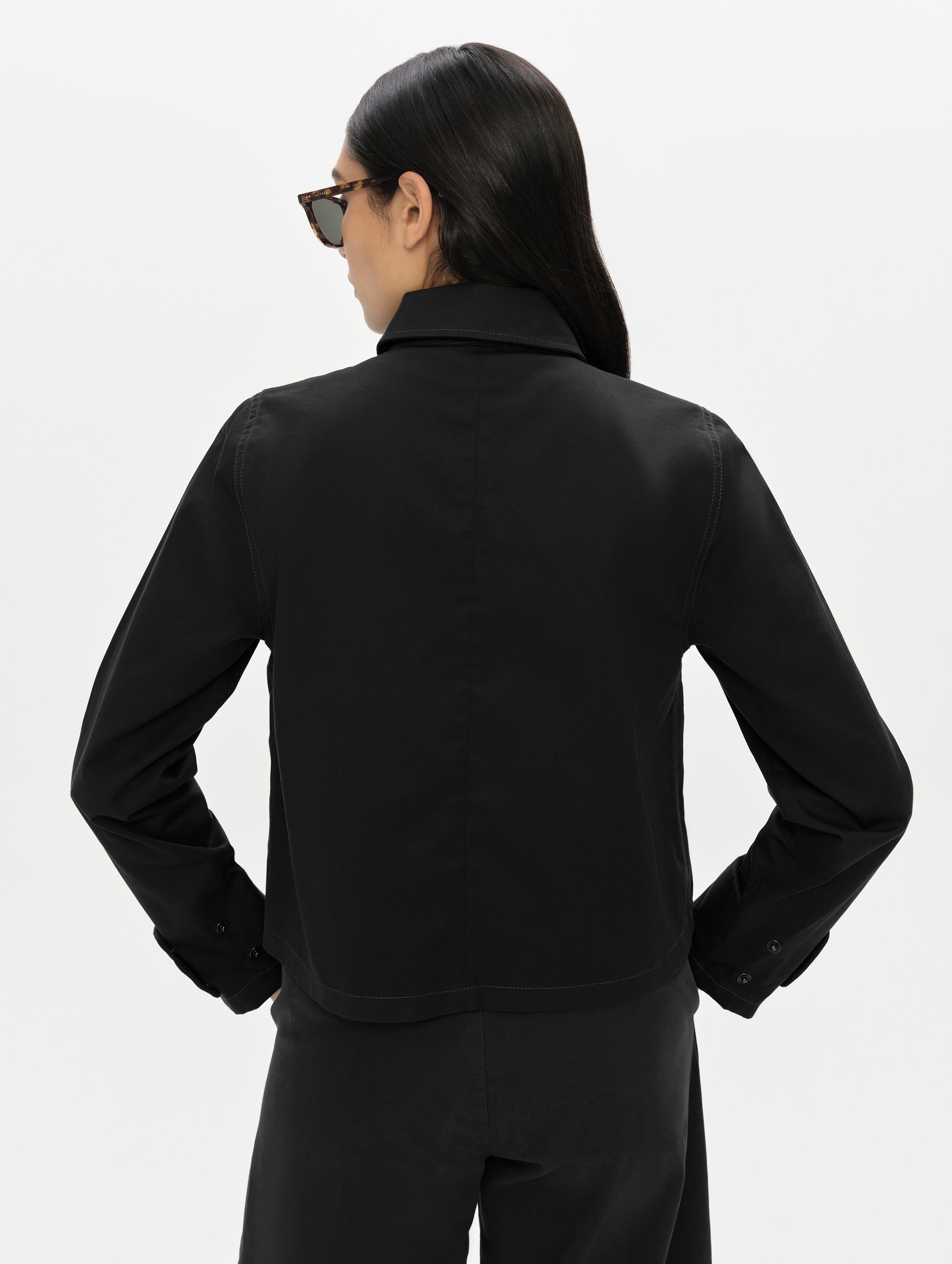 Rear, upper-body view of woman wearing the Raven Waxed Cotton Jacket in Onyx Black from AETHER Apparel.