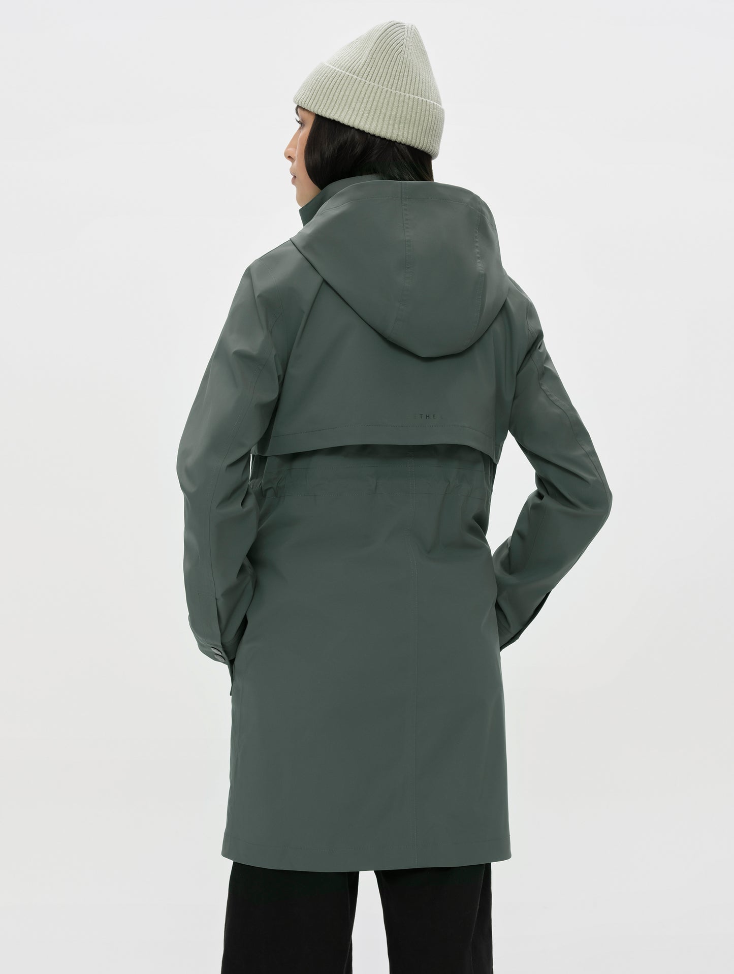 Rear, angled upper-body view of woman wearing the zipped up Wallace Rain Parka in Saville Green from AETHER Apparel.