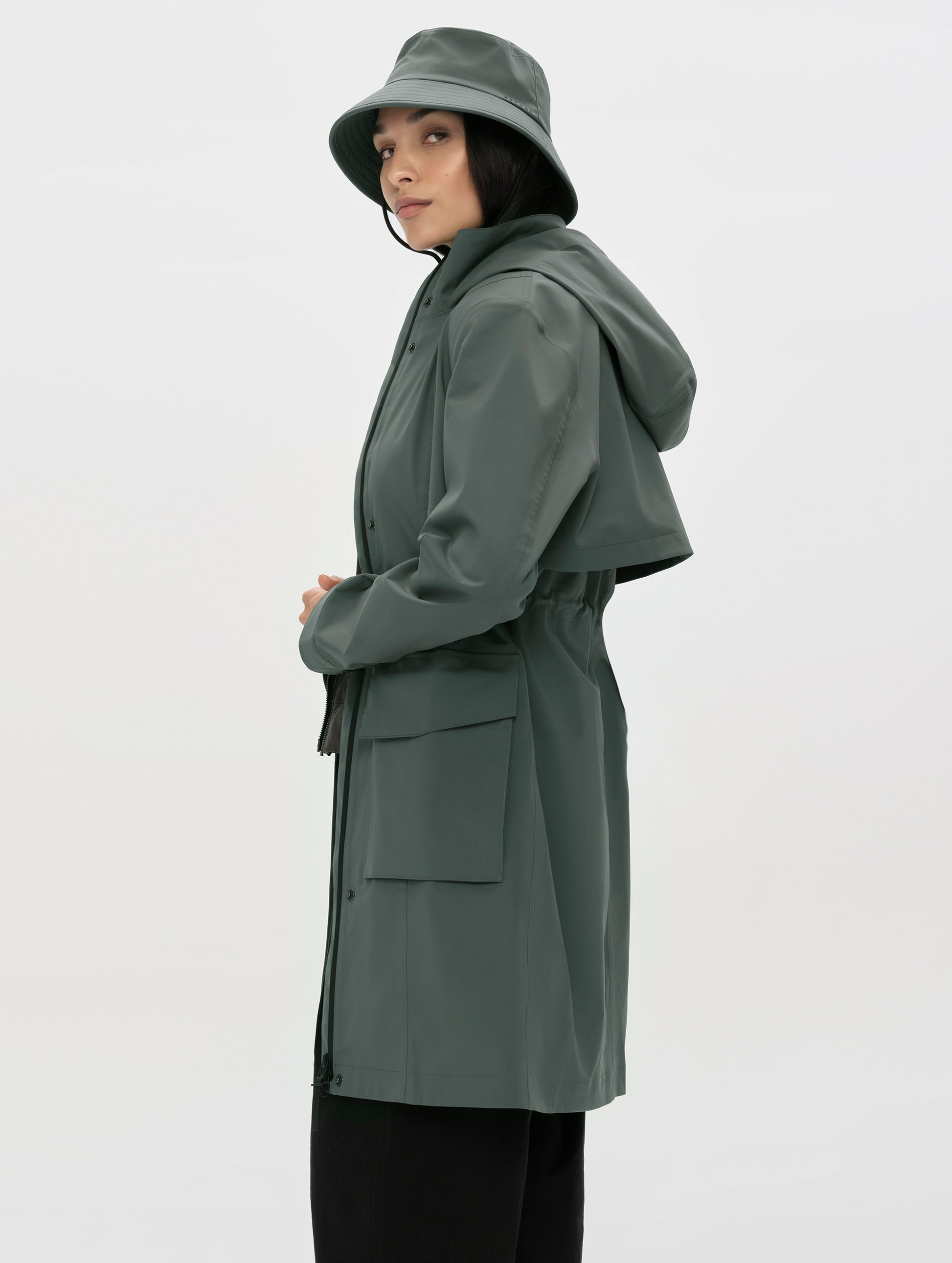 Side profile, upper-body view of woman wearing the Wallace Rain Parka in Saville Green and the Wallace Bucket Hat in Saville Green from AETHER Apparel.