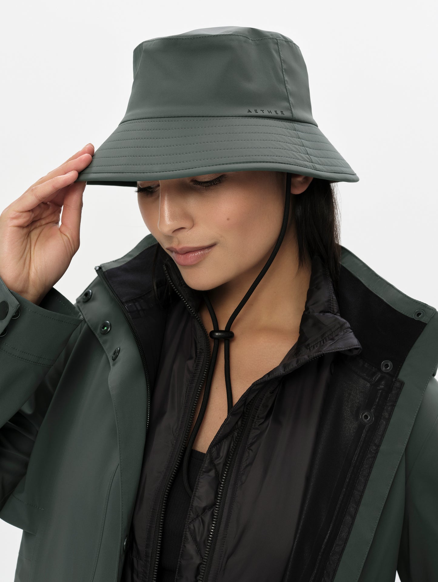 Front right angled view of woman wearing the Wallace Bucket Hat in Saville Green from AETHER Apparel with hand on brim. 