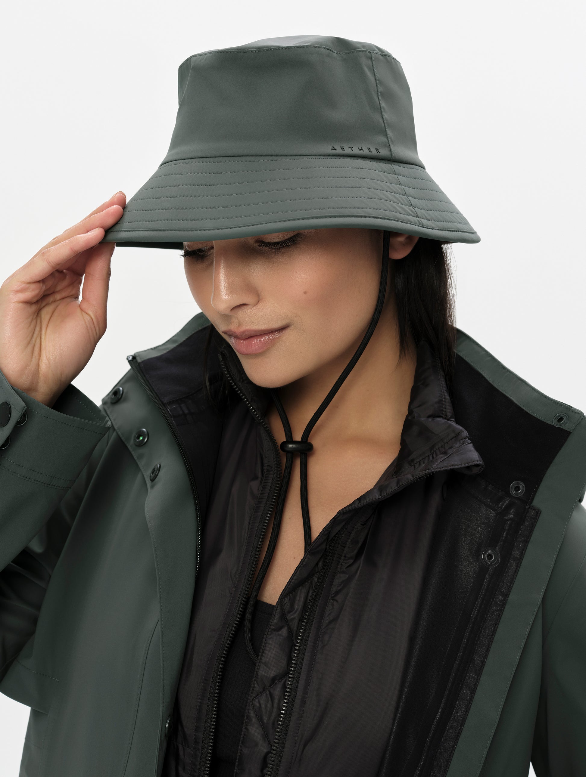 Front right angled view of woman wearing the Wallace Bucket Hat in Saville Green from AETHER Apparel with hand on brim. 