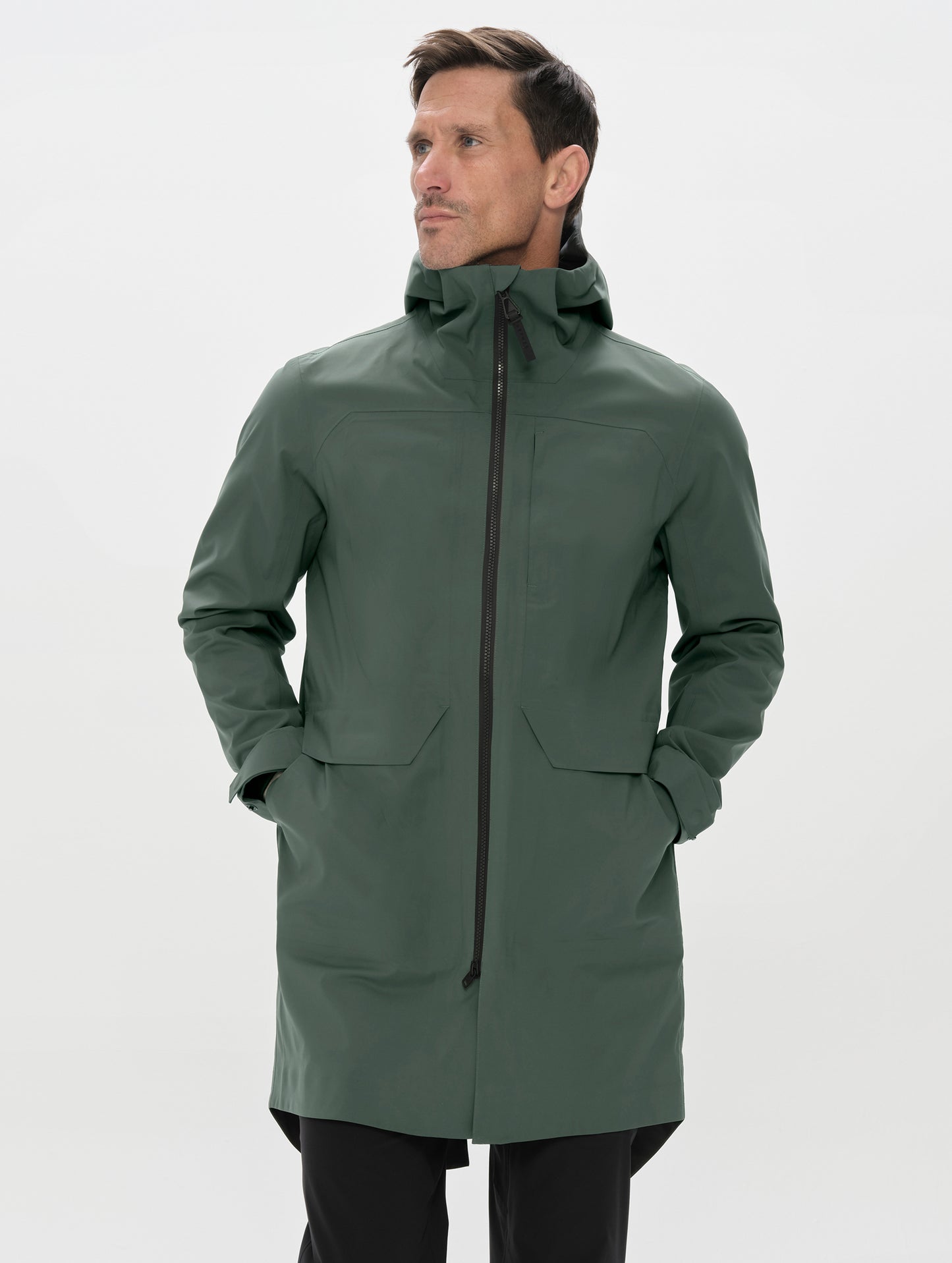 Front upper-body view of man with hands in pockets wearing the Wallace Rain Parka in Saville Green from AETHER Apparel.