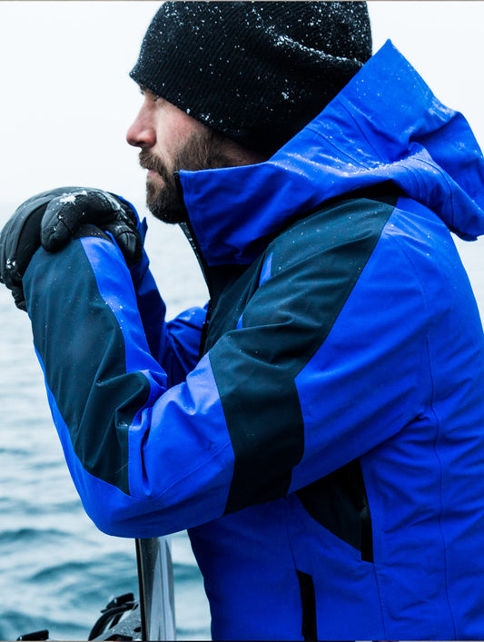 Left side view of man outdoors leaning on  snowboard wearing blue Fusion Shell from AETHER Apparel.