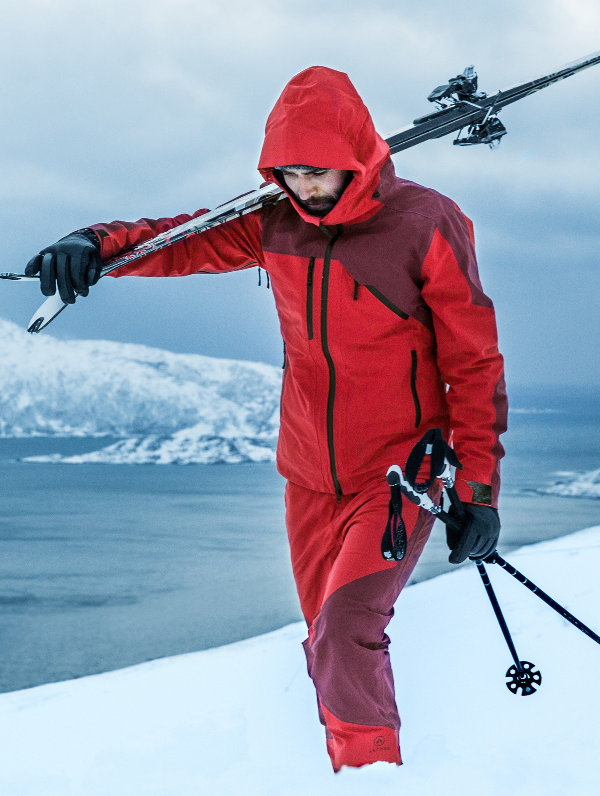 Man outdoors holding skis wearing red Fusion Shell from AETHER Apparel.