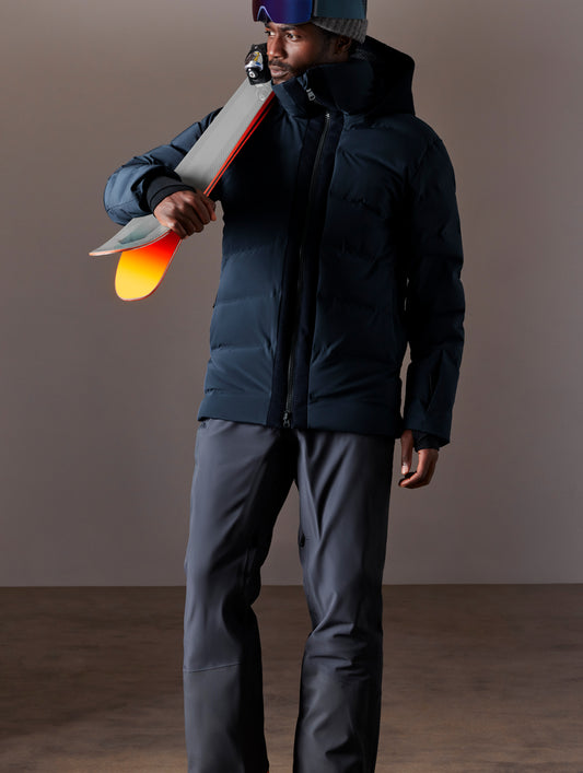 Front full-body view of man holding skis over shoulder wearing Nordic Jacket in Total Eclipse dark blue from AETHER Apparel.